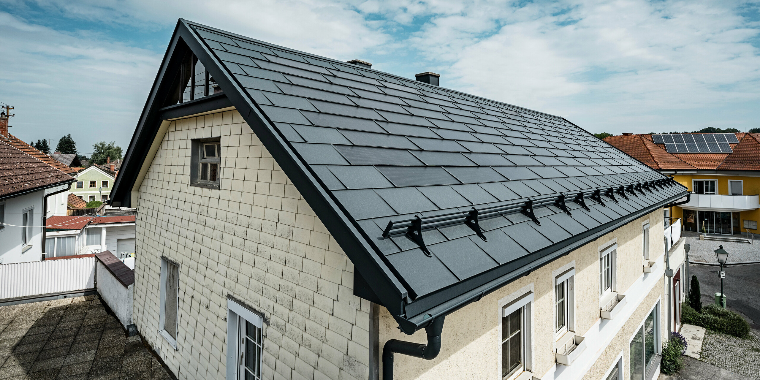 Vogelperspektive auf das Dach eines Einfamilienhauses in Mettmach, Österreich. Das Haus wurde im Zuge einer Sanierung mit PREFA Solardachplatten in Anthrazit ausgestattet. Die strukturierte Oberfläche der Dachplatten harmoniert mit der schlichten Eleganz des Hauses. Ein Schneefangsystem ist am Dachrand angebracht. Im Hintergrund sind weitere Häuser mit traditionellen und solaren Dachlösungen sichtbar, was die moderne, energieeffiziente Dachgestaltung in einem ländlichen Umfeld unterstreicht.