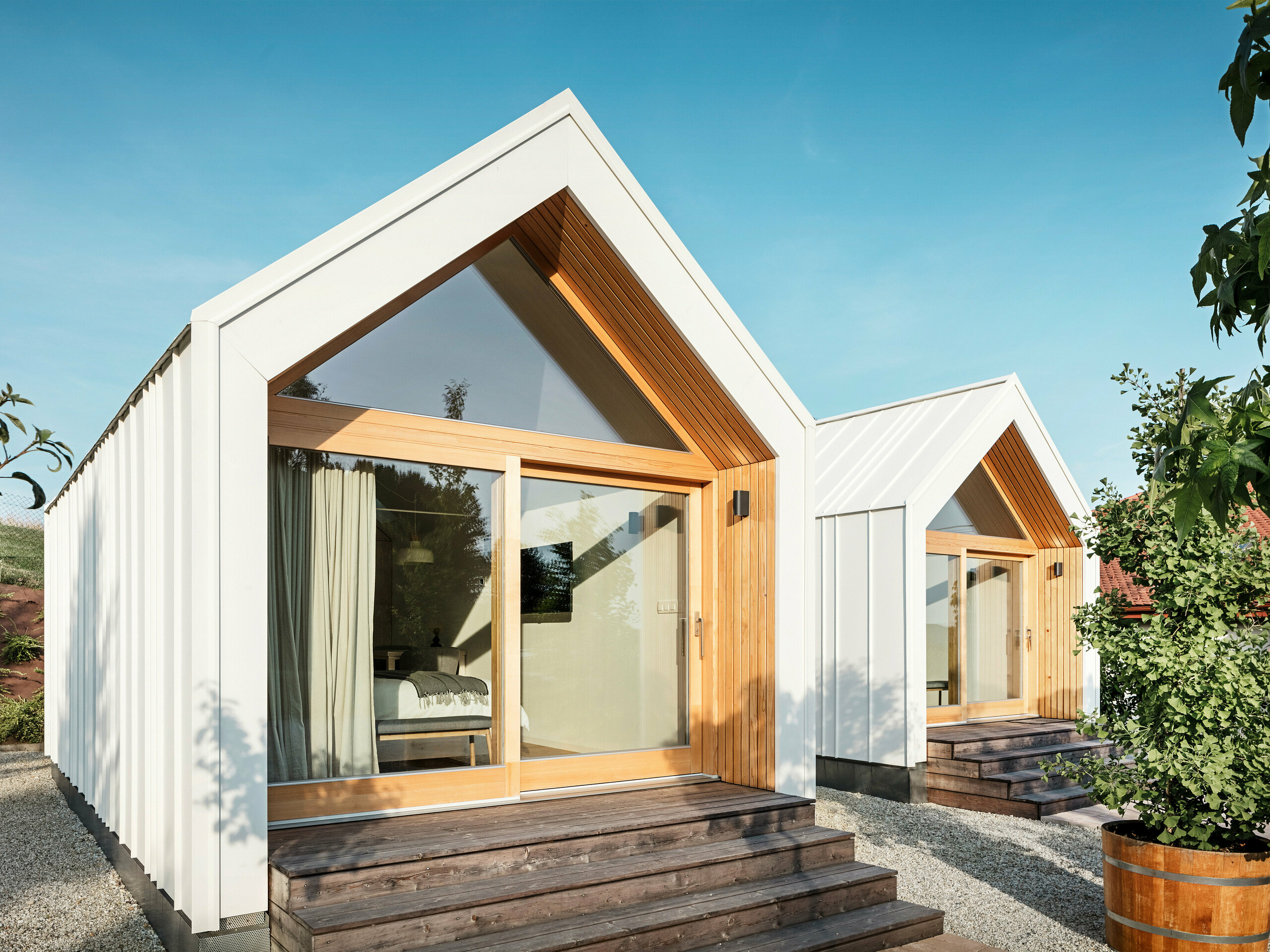 Vue de face des deux petites maisons de la résidence d'hôtes « Pri Momi » à Kančevci, en Slovénie, avec de grandes façades vitrées, conçues avec un toit et un revêtement de façade PREFALZ en blanc pur P.10 et bois naturel. Les micro-maisons minimalistes comportent de larges marches en bois menant aux entrées, offrant une vue directe sur les intérieurs confortables. Les lignes claires et la combinaison de l'aluminium et du bois soulignent le design moderne et accueillant du logement de vacances.