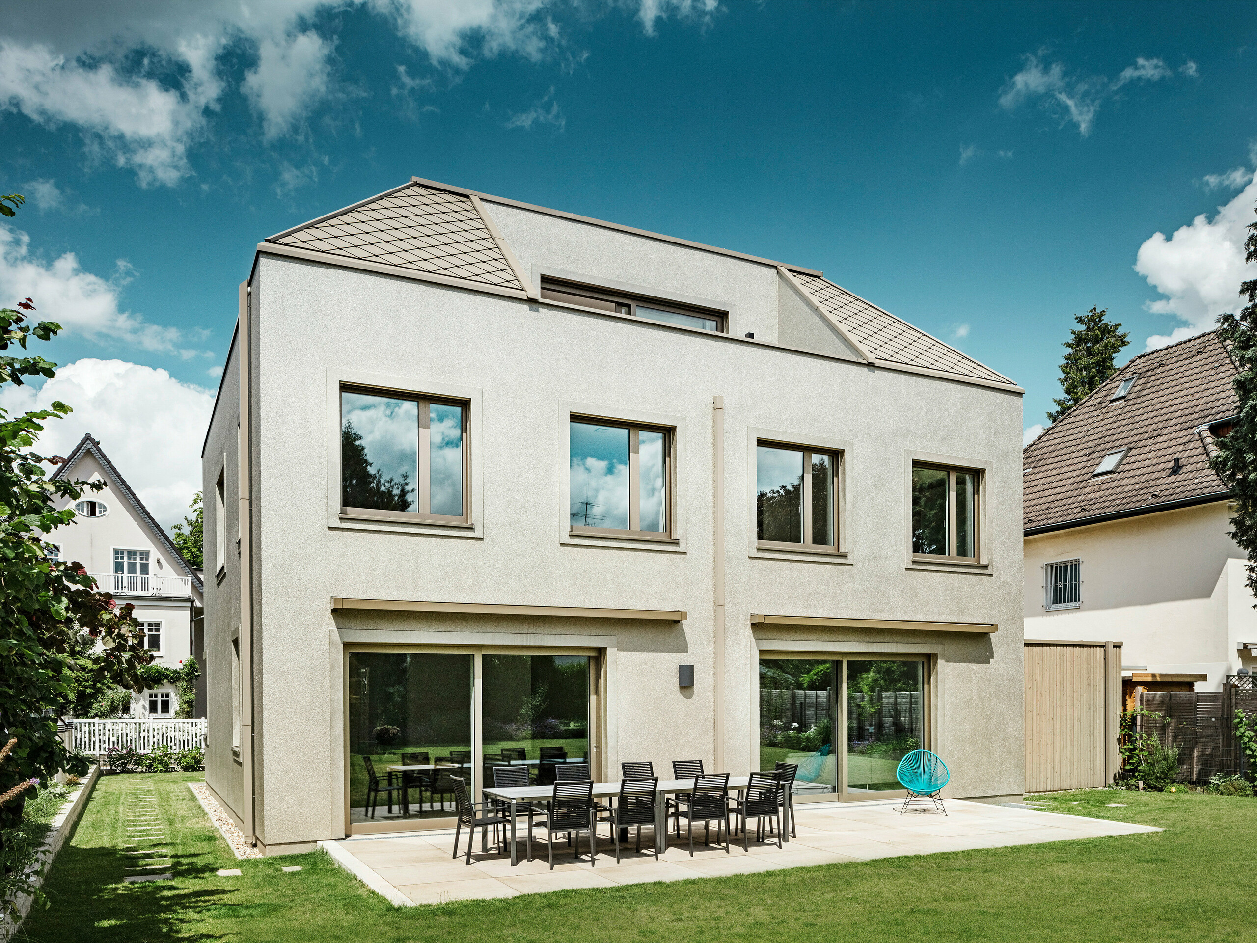 Ein modernes Einfamilienhaus mit Walmdach und bronze-farbener Aluminium-Dach- und Fassadenverkleidung von PREFA. Das Haus befindet sich in einer ruhigen Wohngegend, umgeben von gepflegten Gärten und anderen Einfamilienhäusern. Im Hintergrund sind große Fenster zu sehen, die den Wohnbereich mit viel natürlichem Licht durchfluten. Eine großzügige Terrasse mit Gartenmöbeln lädt zum Entspannen und Verweilen ein. Das Design des Hauses integriert sich harmonisch in die Umgebung, während es durch die moderne Materialwahl von PREFA zeitgemäß und elegant wirkt.