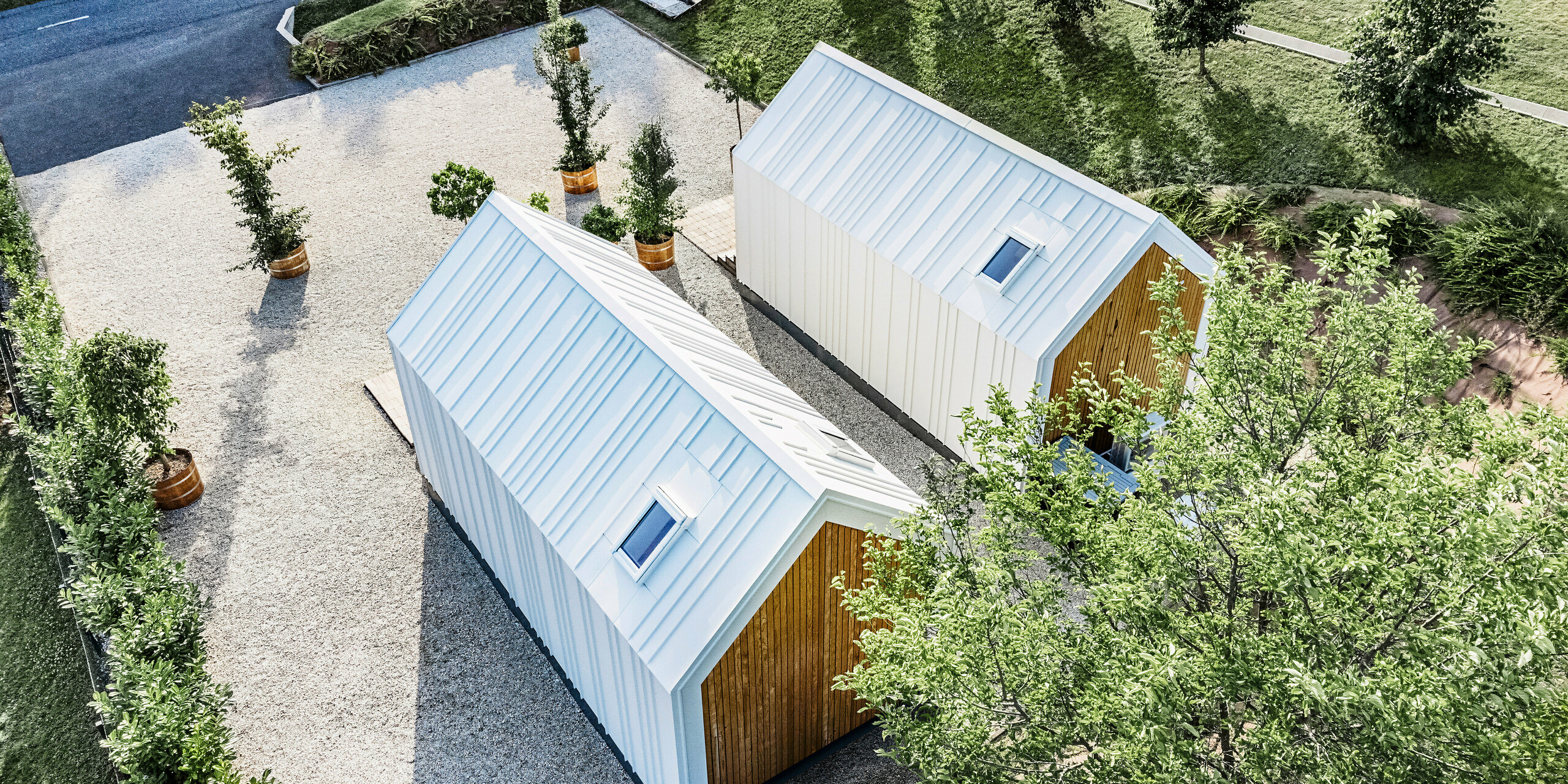 Vue plongeante sur l'arrière des deux petites maisons « Pri Momi » à Kančevci, en Slovénie. Les micro-maisons exclusives sont équipées d'un revêtement de toit et de façade PREFALZ robuste dans la couleur P.10 blanc pur et en bois naturel. L'enveloppe blanche du bâtiment en tôle de haute qualité et durable a été conçue avec un toit à joints debout. L'architecture minimaliste se fond harmonieusement dans l'espace vert environnant et les allées pavées. La photo montre également la disposition légèrement décalée des bâtiments, qui ont été construits à ossature bois.