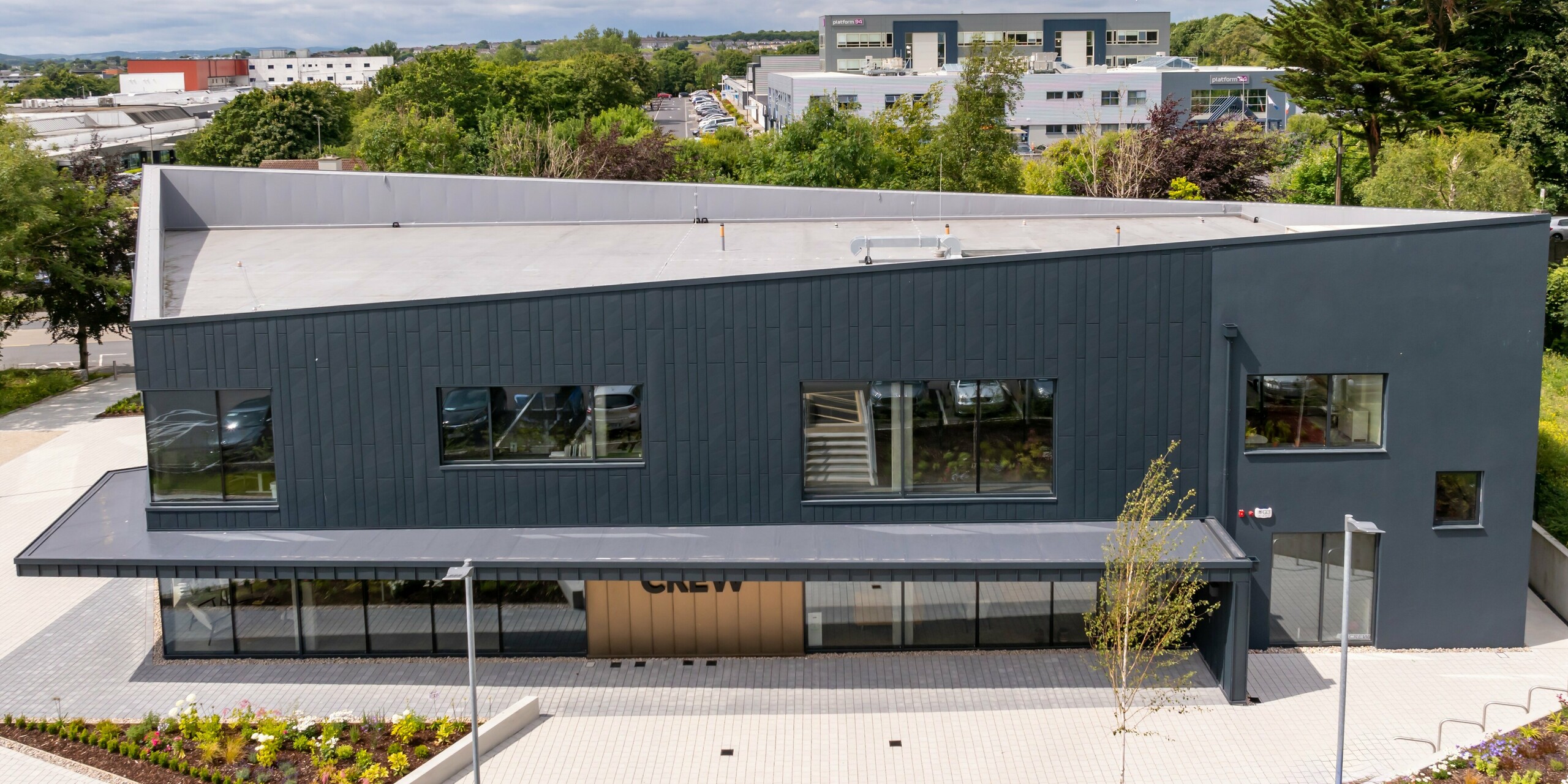 Vue aérienne du bâtiment CREW à Galway, en Irlande, avec une façade moderne composée de panneaux de façade PREFA Siding.X et PREFALZ en anthracite P.10. Les lignes de toit en pente et les zones de fenêtres clairement structurées soulignent le design innovant du bâtiment. Le bardage en aluminium de couleur anthracite s'harmonise avec l'environnement et confère au bâtiment un aspect contemporain et de grande qualité.