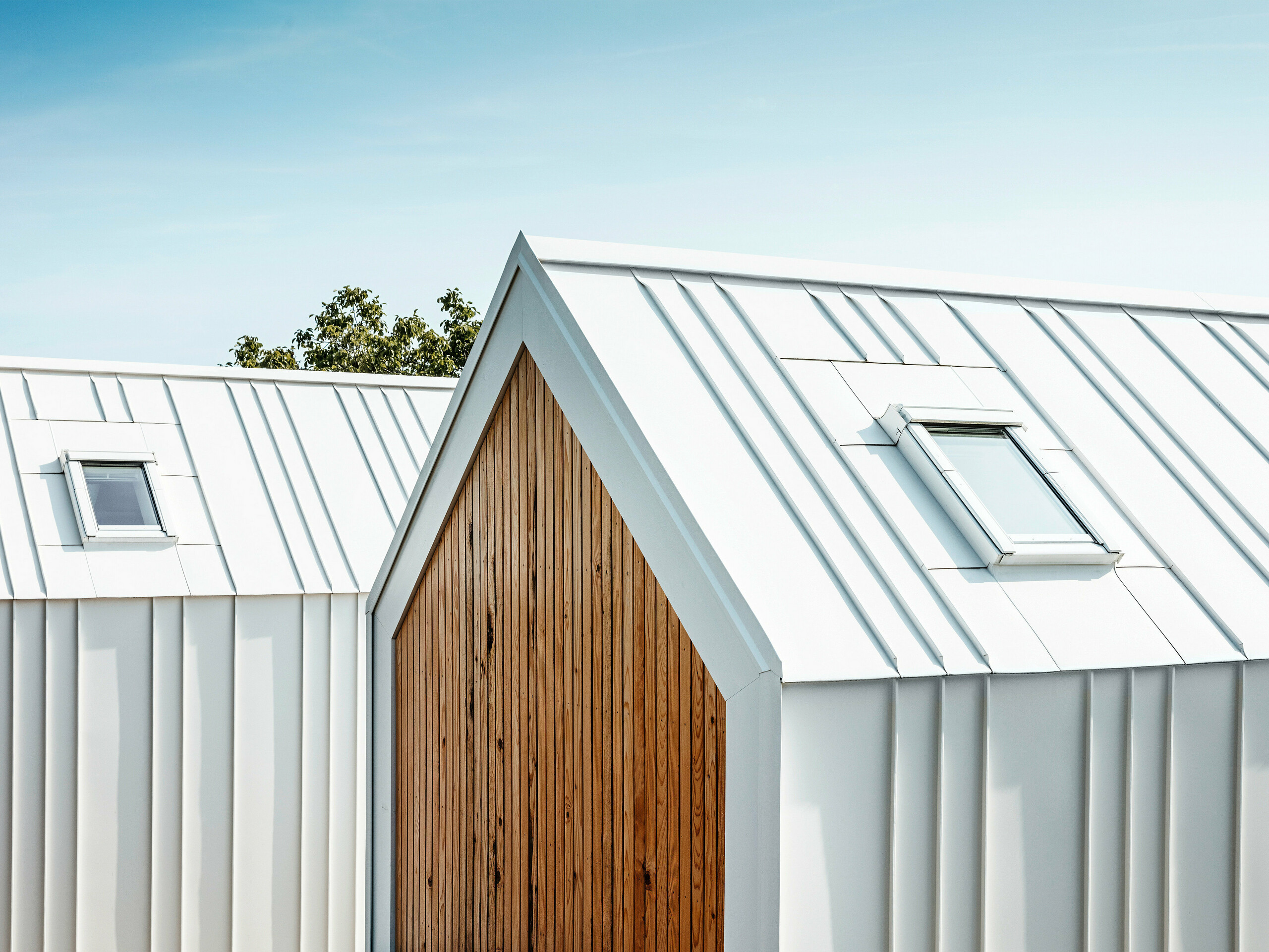 Vue détaillée revêtement de toiture et du de façade PREFALZ blanc des petites maisons « Pri Momi » à Kančevci, Slovénie. Les bâtiments sont conçus dans une combinaison moderne d'aluminium de haute qualité en blanc pur P.10 et de bois naturel. La photo montre les plis exécutés avec précision et la transition élégante du toit à la façade, qui a été réalisée sans aucune interruption visible des joints verticaux. Le choix harmonieux des matériaux souligne le design minimaliste de l'hébergement exclusif.