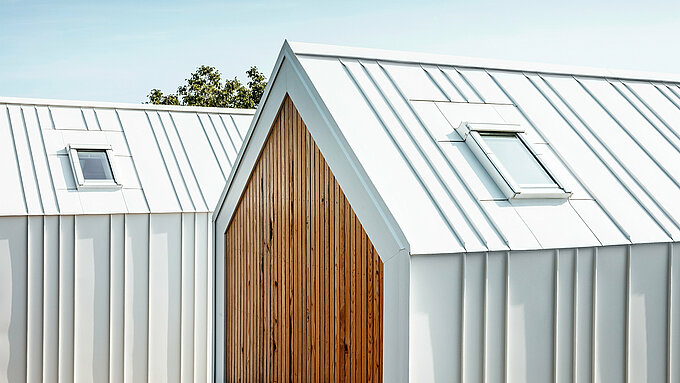 Vue détaillée revêtement de toiture et du de façade PREFALZ blanc des petites maisons « Pri Momi » à Kančevci, Slovénie. Les bâtiments sont conçus dans une combinaison moderne d'aluminium de haute qualité en blanc pur P.10 et de bois naturel. La photo montre les plis exécutés avec précision et la transition élégante du toit à la façade, qui a été réalisée sans aucune interruption visible des joints verticaux. Le choix harmonieux des matériaux souligne le design minimaliste de l'hébergement exclusif.