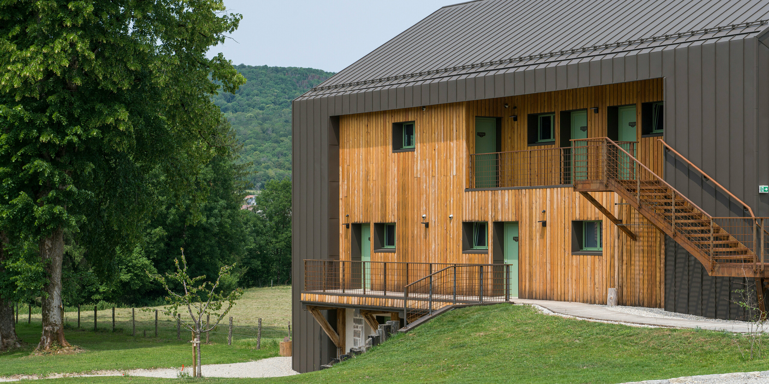 Perspective latérale de l'Hôtel en Tilleul à Pont du Navoy (France). Le bâtiment moderne présente une enveloppe extérieure harmonieuse en bois et en aluminium. PREFALZ dans la couleur P.10 brun a été utilisé pour les éléments en aluminium du toit et des bords de la façade. L'aluminium brun s'intègre parfaitement au paysage naturel avec de nombreuses collines et arbres. Le matériau est également le complément idéal à la façade en bois clair. De nombreuses fenêtres et un escalier avec rampe métallique menant aux étages supérieurs agrémentent la face visible du bâtiment.