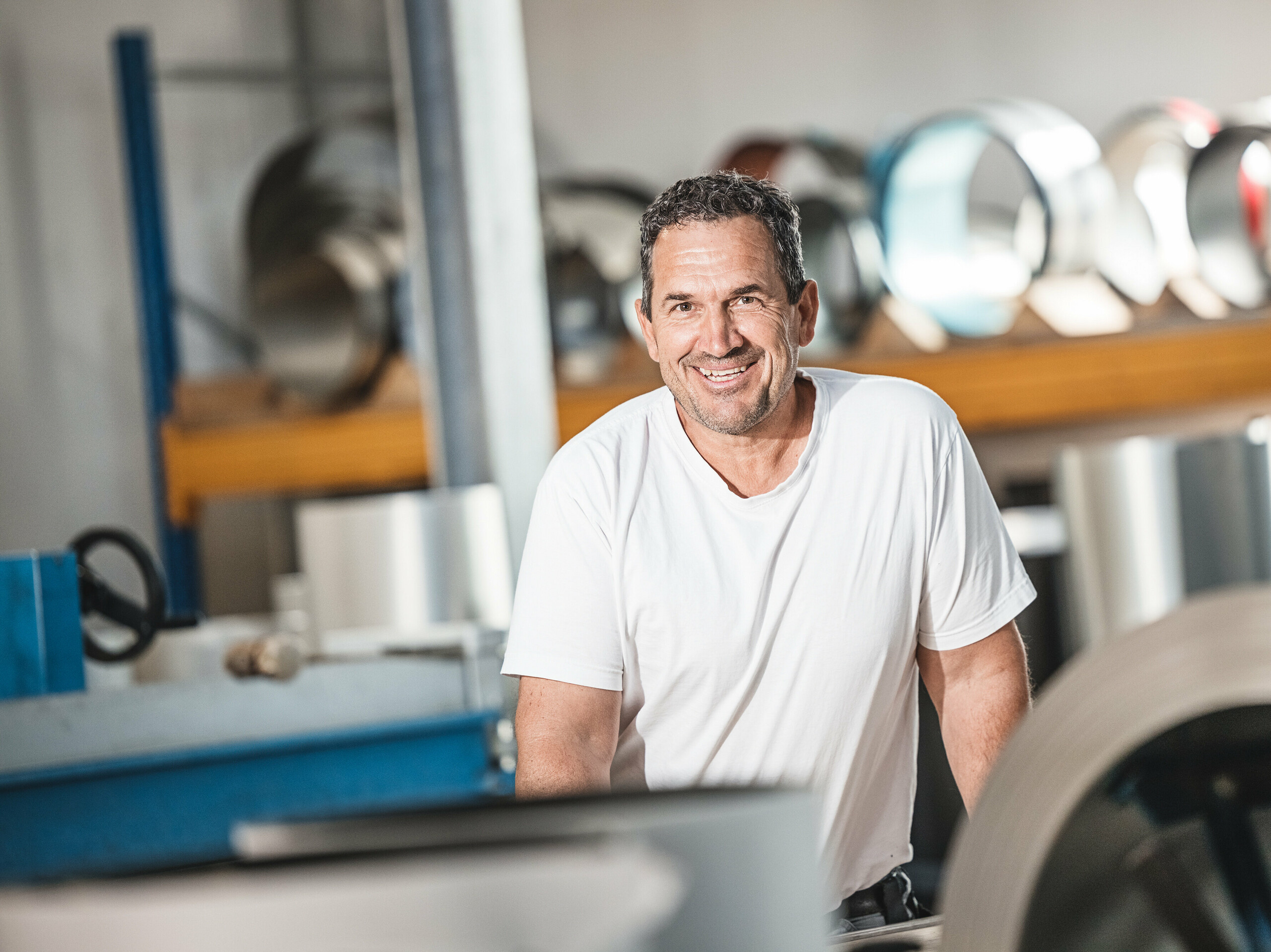 Oskar Urban de l'entreprise de toiture Urban dans son atelier. L'installateur PREFA expérimenté se tient souriant devant un fond de rouleaux et de machines en aluminium qui soulignent la haute qualité et la précision de son travail avec les produits PREFA. Son expertise contribue de manière significative à la mise en œuvre réussie de projets tels que le complexe immobilier du site Schmucker à Utting am Ammersee, où se combinent architecture moderne et matériaux durables.