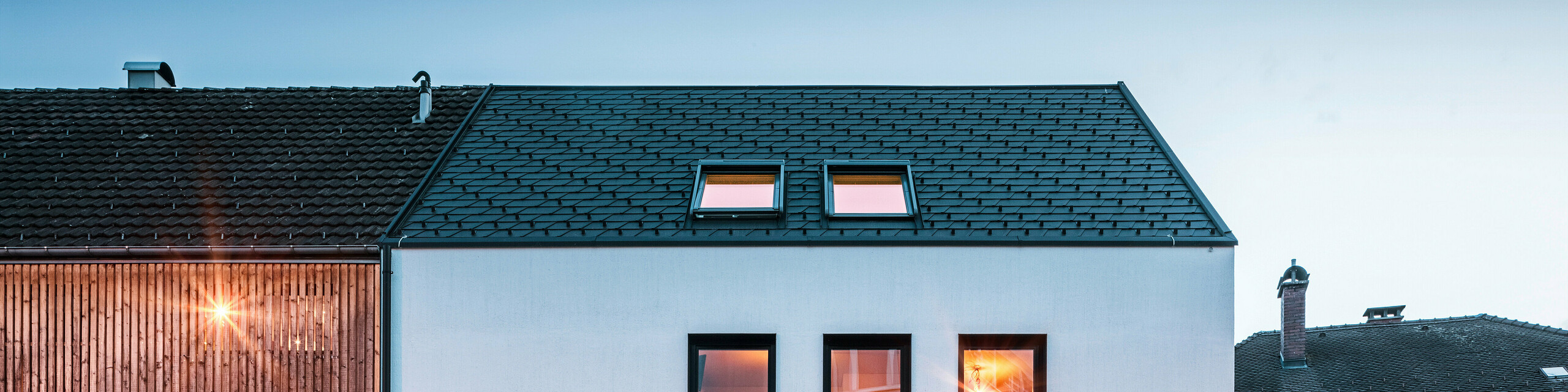 Façade moderne d'un immeuble d'habitation avec bardeaux PREFA DS19 et Prefalz en noir au bord du lac de Constance en Autriche. Le bâtiment est clairement divisé en deux, avec une extension en bois sur la gauche et une partie principale crépie sur la droite. Le toit présente la couverture en bardeaux foncés caractéristique et il y a plusieurs fenêtres rectangulaires qui se reflètent dans la chaude lumière du soir.