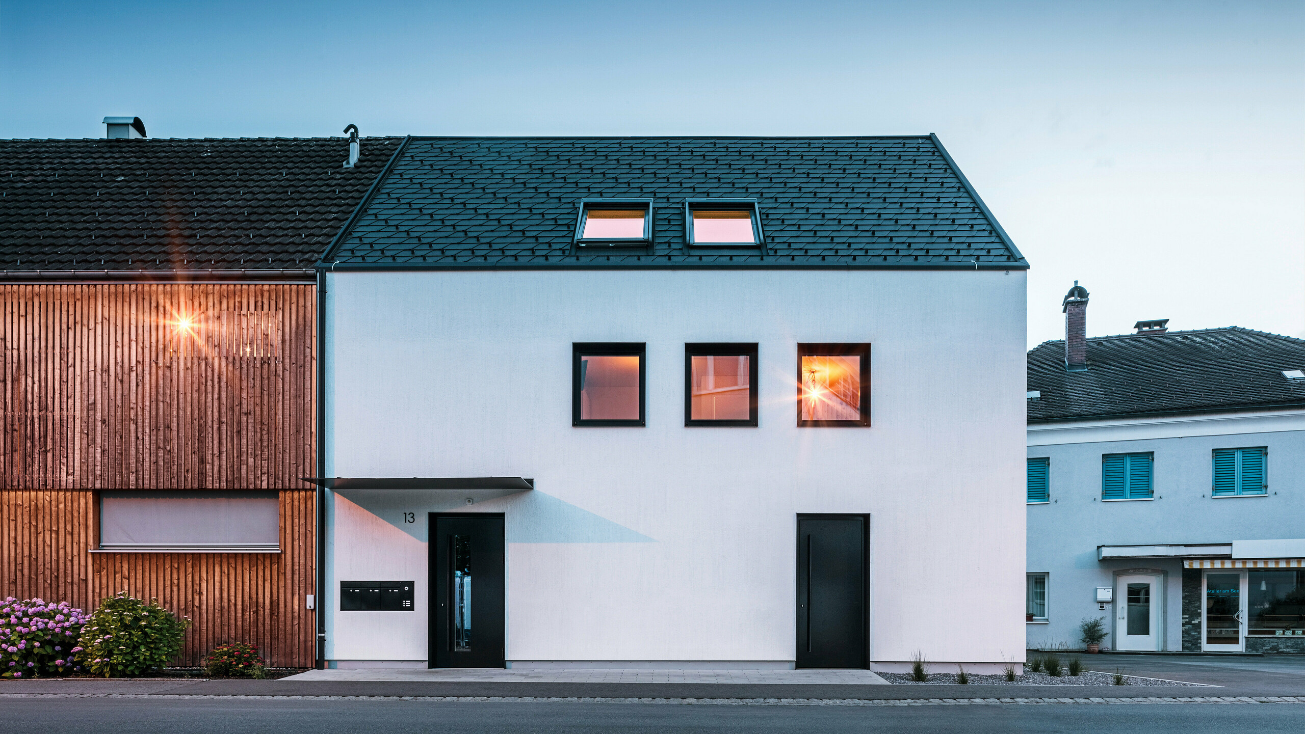 Façade moderne d'un immeuble d'habitation avec bardeaux PREFA DS19 et Prefalz en noir au bord du lac de Constance en Autriche. Le bâtiment est clairement divisé en deux, avec une extension en bois sur la gauche et une partie principale crépie sur la droite. Le toit présente la couverture en bardeaux foncés caractéristique et il y a plusieurs fenêtres rectangulaires qui se reflètent dans la chaude lumière du soir.