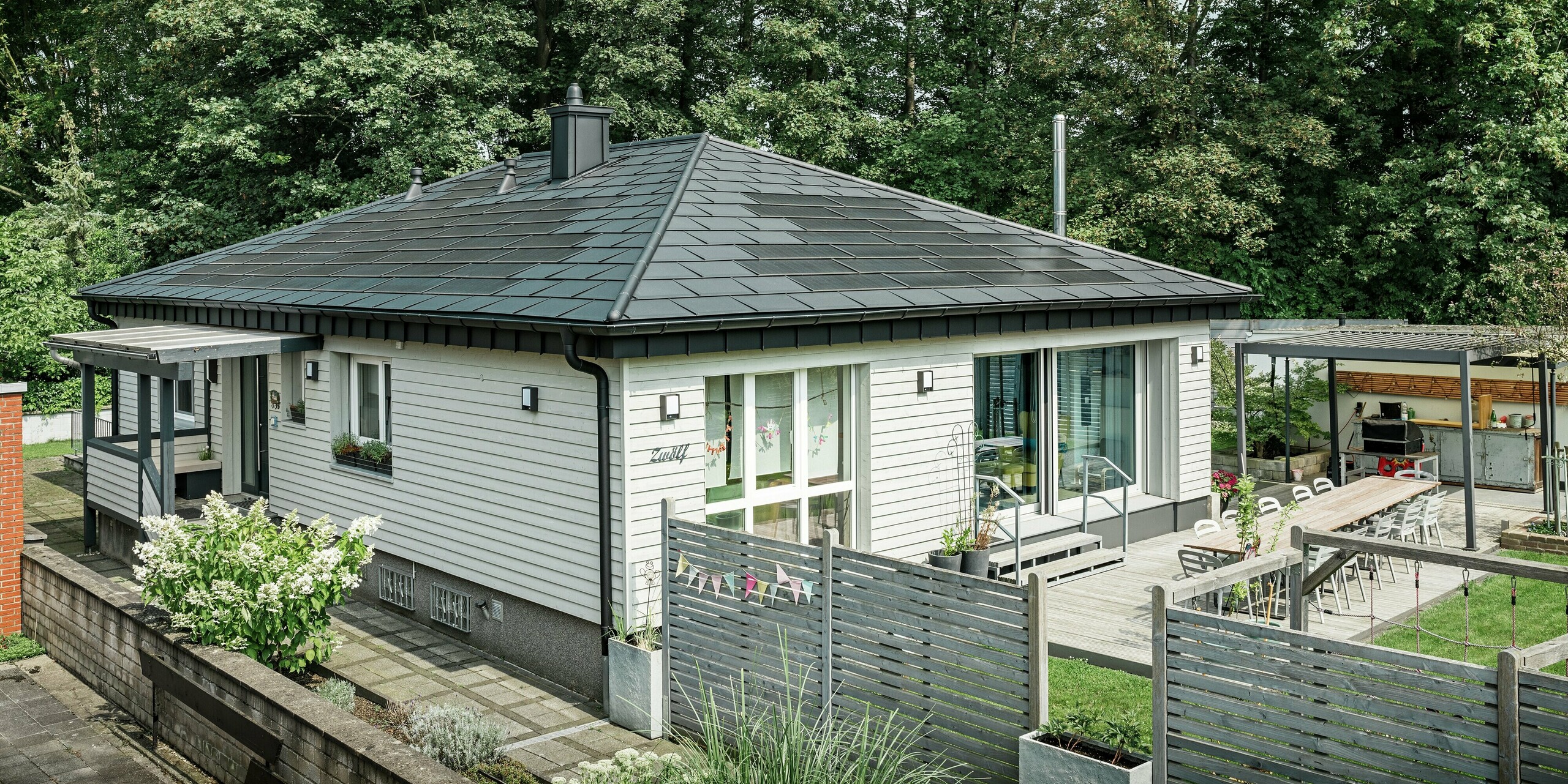 Un bungalow à Rommerskirchen (Allemagne) après rénovation avec un élégant toit en aluminium PREFA. La combinaison innovante des tuiles solaires PREFA et des R.16 en P.10 noir offre une protection durable tout en produisant de l'énergie en même temps. La gouttière et le tuyau de descente sont parfaitement intégrés et assurent un drainage efficace du toit dans une conception de haute qualité. L'accès à la maison familiale est sécurisé par une clôture en bois. Entre les éléments de clôture se trouvent des lits bas qui offrent une vue sur le jardin et la terrasse.