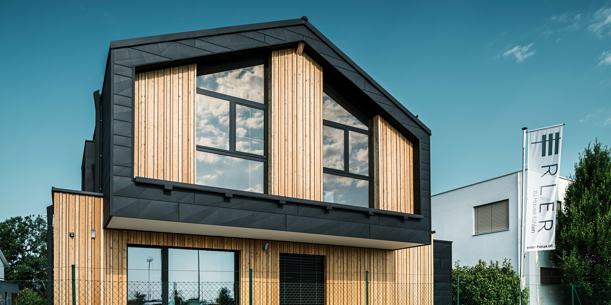 Façade de la maison modèle Autark dans le centre de maisons préfabriquées Blaue Lagune, Vösendorf (AT). La façade combine des éléments verticaux en bois avec les panneaux PREFA Siding.X en aluminium noir. De grandes surfaces vitrées sur le pignon reflètent le ciel et donnent à la maison un aspect moderne et inondé de lumière. Le design met l'accent sur l'harmonie entre le bois, le verre et l'aluminium pour une architecture contemporaine et durable.