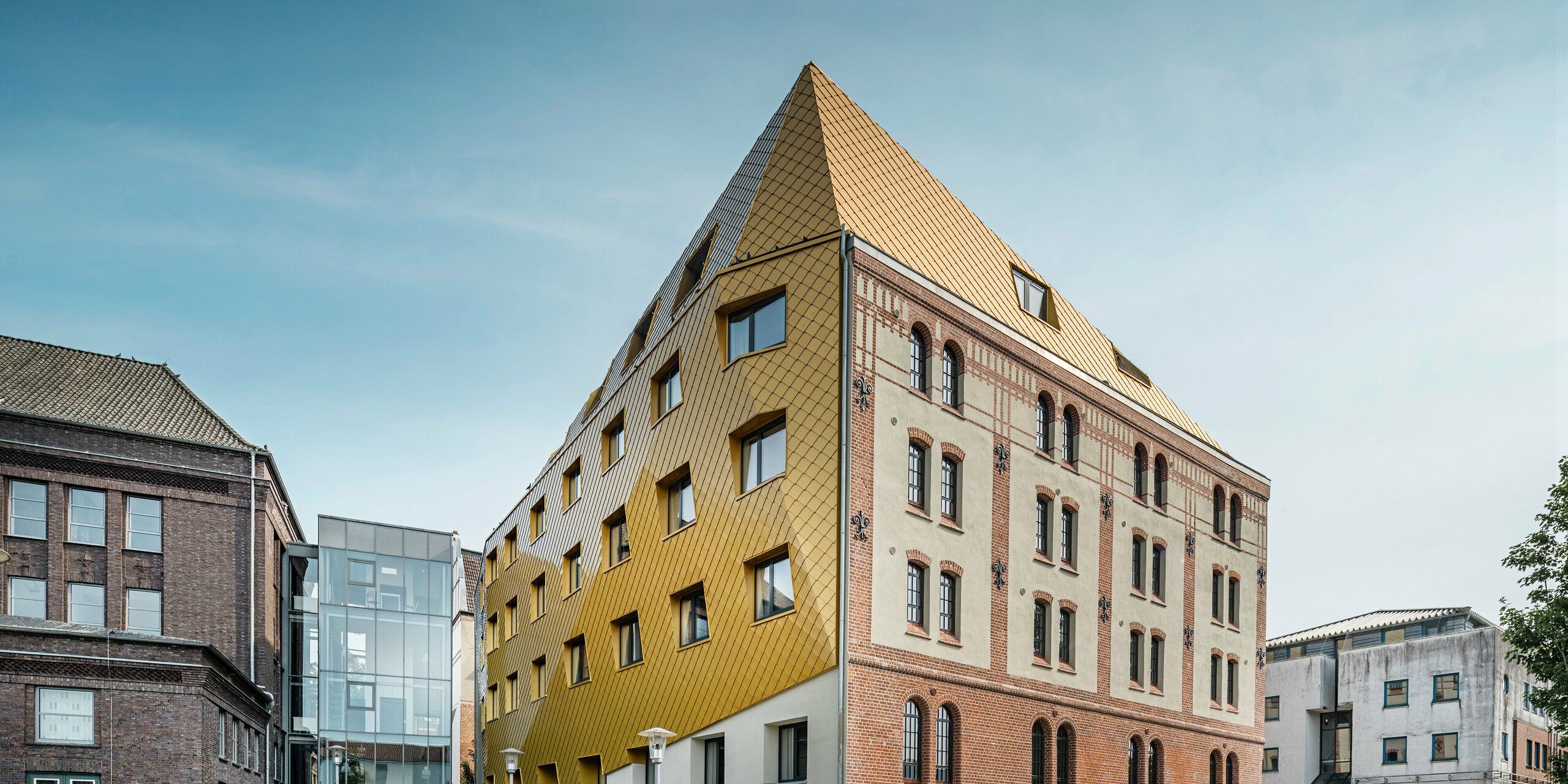 Bodenaufnahme auf "The Station" in Kiel. Das Studentenwohnheim ist schräg von der Seite zu sehen. Auf dem Bild wird der eklatante Kontrast zwischen der Aluminiumfassade aus PREFA Dach- und Wandrauten 29 × 29 in Mayagold auf der linken Seite und der denkmalgeschützten Putz- und Ziegelfassade auf der rechten Seite deutlich.