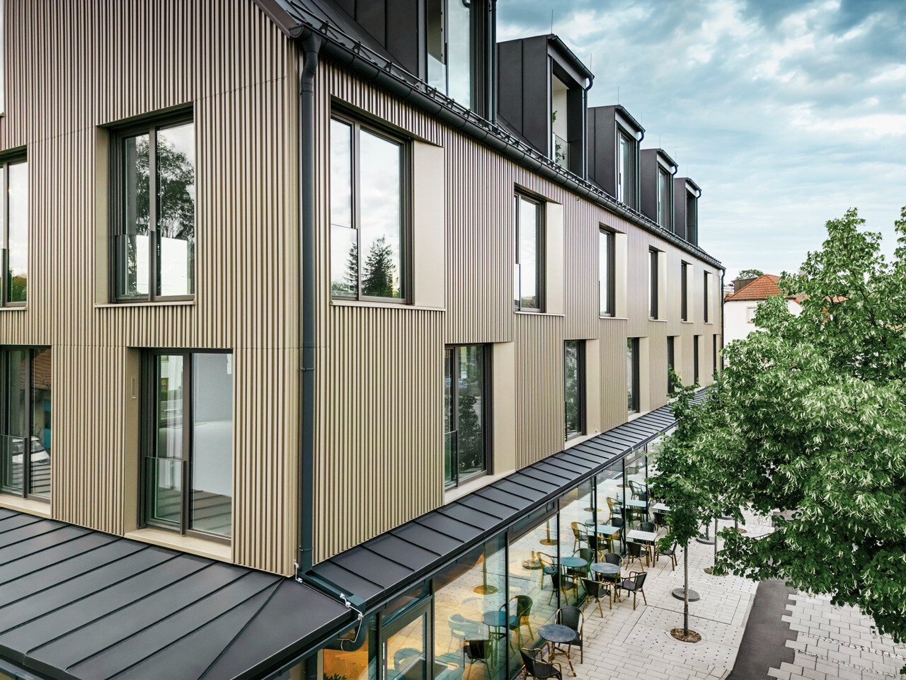 La photo montre un bâtiment moderne à Feldkirchen, conçu par l'architecte Stefan Heigl. La façade est en Prefalz de couleur bronze et le toit à pignon est recouvert de Prefalz de couleur gris noir. Les grandes fenêtres apportent beaucoup de lumière à l'intérieur, tandis que le rez-de-chaussée vitré offre un coin repas accueillant. Le choix des matériaux de haute qualité et le design élégant confèrent au bâtiment un aspect contemporain et élégant.