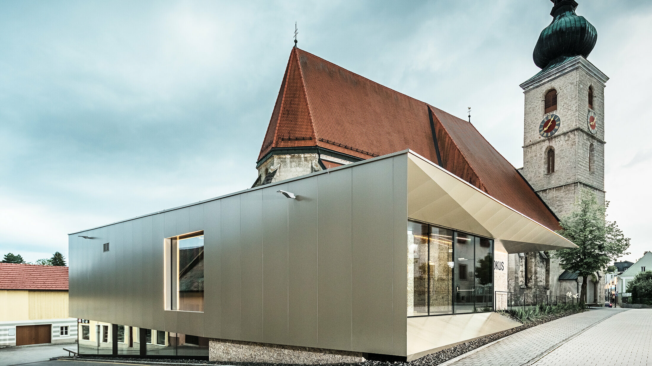 La façade marquante du centre paroissial de Sierning avec ses panneaux composites couleur de bronze s’étend autour de la petite église en arrière-plan