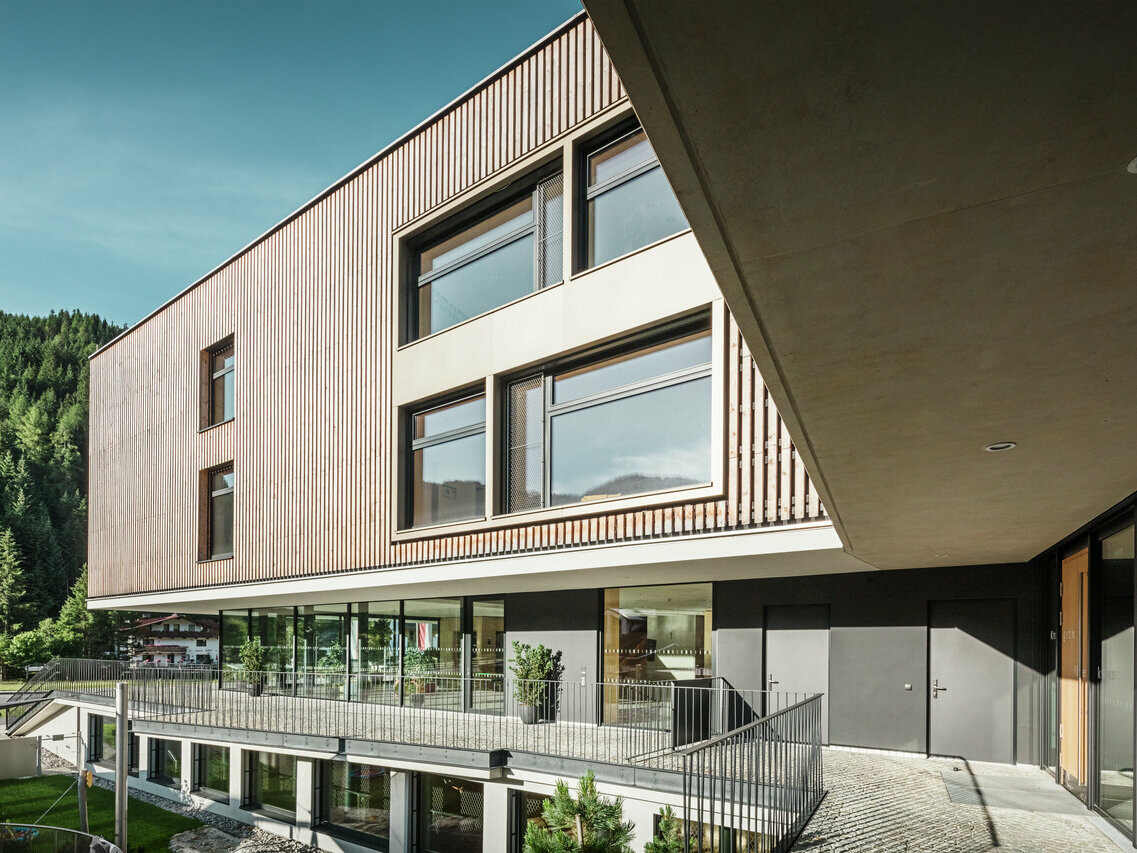 Architecture moderne d'un bâtiment de plusieurs étages combinant une façade en bois et en crépi, intégrée dans un environnement alpin avec des collines boisées en arrière-plan. Le bâtiment dispose de grandes fenêtres qui laissent entrer beaucoup de lumière à l'intérieur et d'une terrasse couverte qui accentue l'entrée.