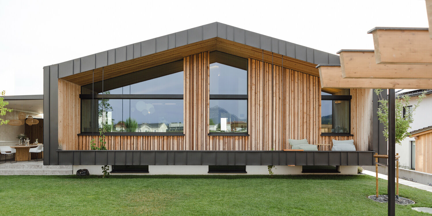 Modernes Einfamilienhaus in Kirchham, Österreich, mit einer beeindruckenden Fassade aus naturbelassenem Holz und großzügigen Glasfronten. Das Dach und Teile der Fassade sind mit hochwertigem PREFALZ in P.10 Anthrazit von PREFA veredelt, was einen eleganten Kontrast zur organischen Holzstruktur schafft. Die klaren Linien und das zeitgenössische Design spiegeln das innovative Konzept von PREFA Aluminiumprodukte wider, während die umlaufende Fensterfront einen ungehinderten Blick auf die gepflegten Grünflächen und den Pool ermöglicht sowie das Innere mit natürlichem Licht durchflutet.