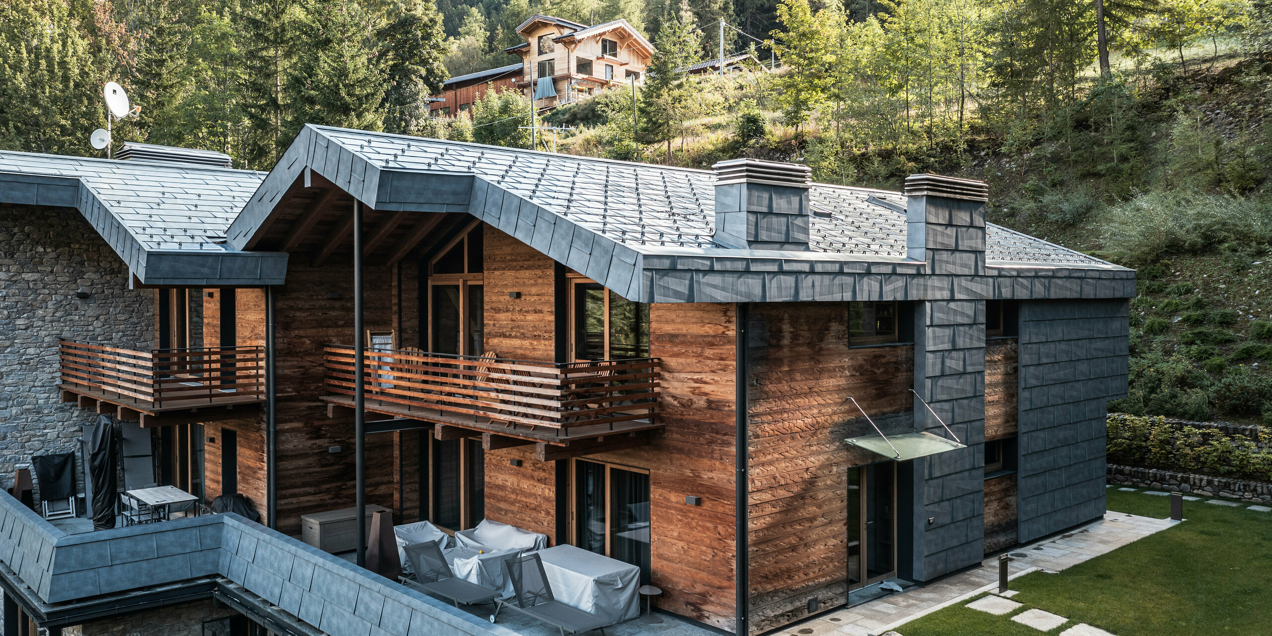 Detailaufnahme einer modernen Villa in Courmayeur, die sich durch die Verwendung von Holz, Stein und Blech an der Fassade auszeichnet. Die PREFA FX.12 Dach- und Fassadenpaneele in P.10 Steingrau bilden einen optischen Höhepunkt des modernen architektonischen Gebäudes. Die Villa verfügt über mehrere Balkone und Terrassen, die einen ruhigen Rückzugsort im Freien mit Blick auf die dichte Waldlandschaft bieten. Architektonische Merkmale wie die scharfen Kanten des Daches und das robuste Aluminium ergänzen den rustikalen Charakter der Umgebung.