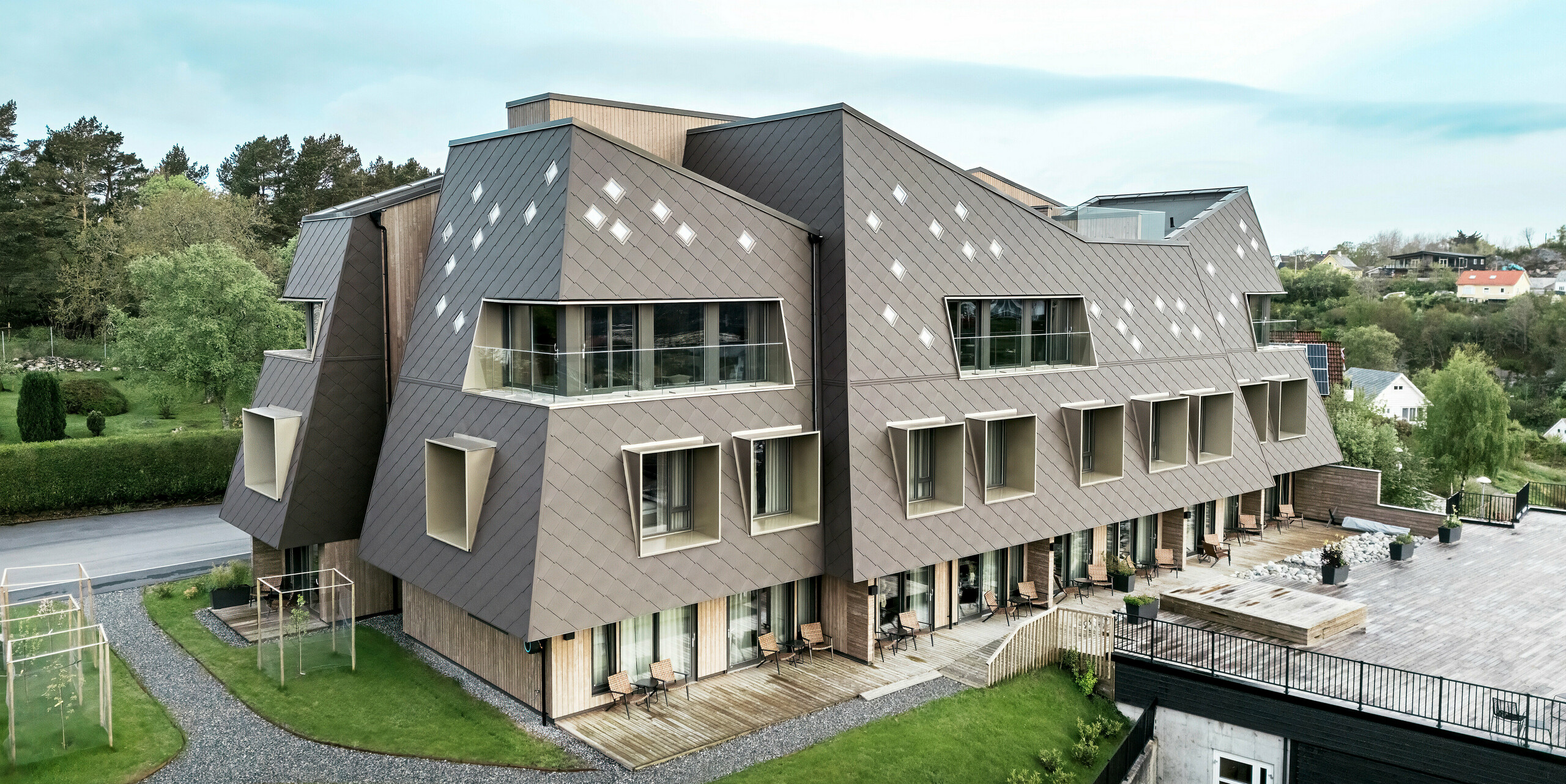 Vue extérieure de la maison Beckerwyc à Bekkjarvik, en Norvège, avec un accent sur les espaces extérieurs bien entretenus et la façade saisissante composée de losanges PREFA 44 × 44 en brun noisette. L'architecture sculpturale du bâtiment, combinée à une terrasse et des balcons spacieux, crée une connexion harmonieuse entre les espaces intérieurs et extérieurs. Le design futuriste et les couleurs naturelles s'intègrent bien dans le paysage environnant.