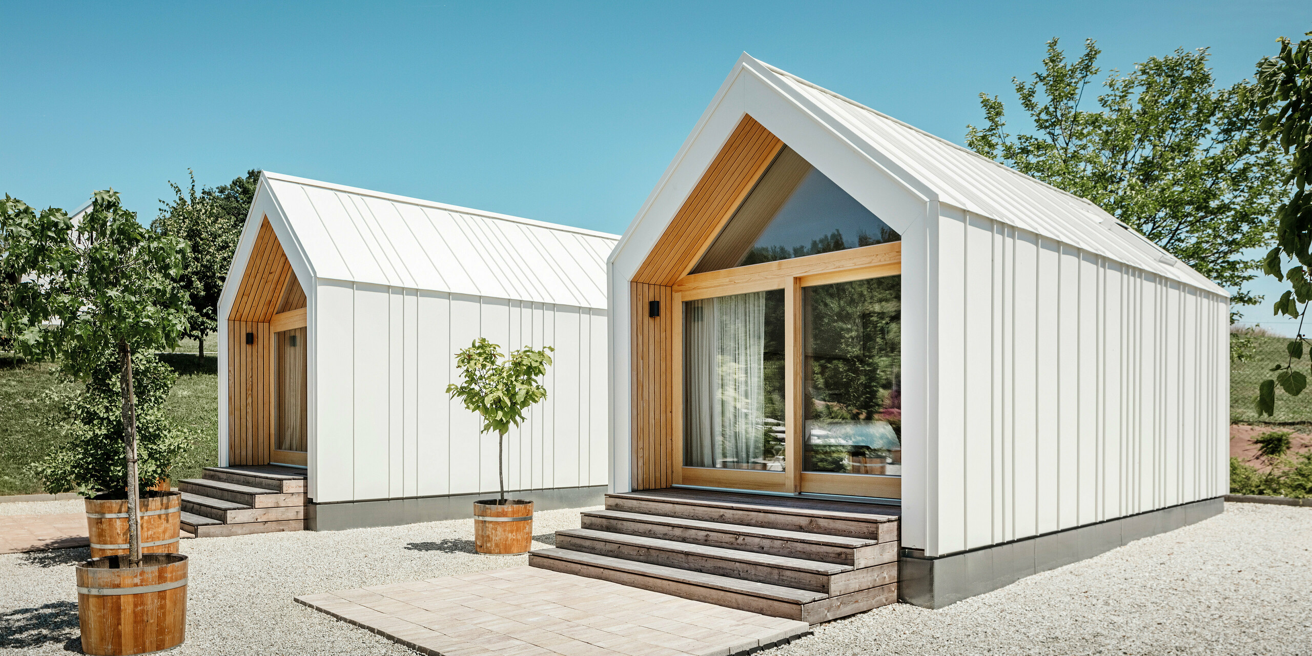 Deux petites maisons blanches dans l'idylle de vacances « Pri Momi » à Kančevci, en Slovénie, conçues avec un revêtement de toit et de façade PREFALZ en blanc pur P.10 et en bois naturel. L'architecture minimaliste des bâtiments est soulignée par l'élégant mélange de matériaux aluminium et bois. Les grandes façades vitrées aux entrées ainsi que les marches en bois et les arbres environnants créent une atmosphère invitante et harmonieuse. L'hébergement exclusif est situé au milieu d'un paysage pittoresque et est utilisé spécifiquement pour les célébrations.