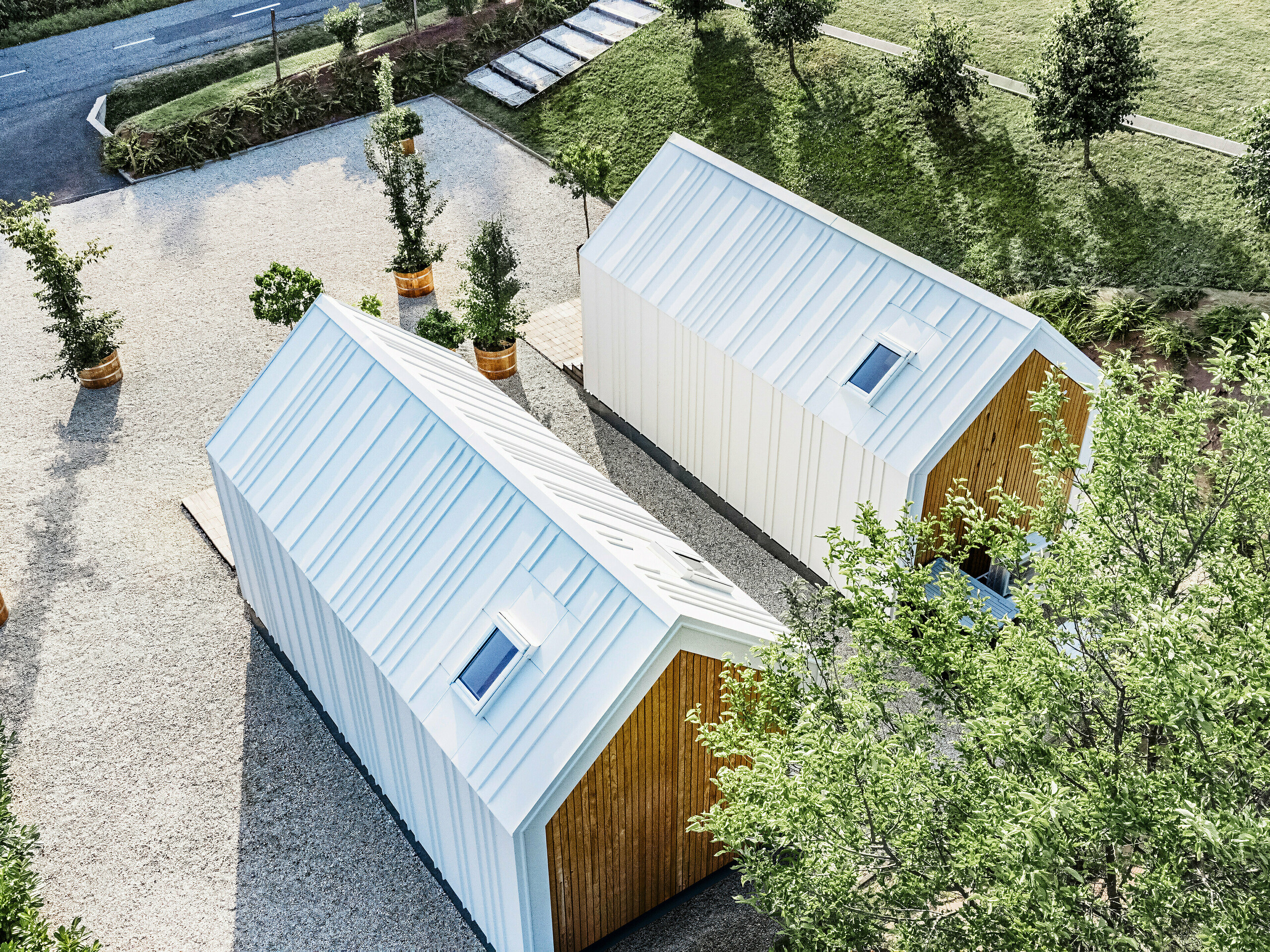 Vue plongeante sur l'arrière des deux petites maisons « Pri Momi » à Kančevci, en Slovénie. Les micro-maisons exclusives sont équipées d'un revêtement de toit et de façade PREFALZ robuste dans la couleur P.10 blanc pur et en bois naturel. L'enveloppe blanche du bâtiment en tôle de haute qualité et durable a été conçue avec un toit à joints debout. L'architecture minimaliste se fond harmonieusement dans l'espace vert environnant et les allées pavées. La photo montre également la disposition légèrement décalée des bâtiments, qui ont été construits à ossature bois.