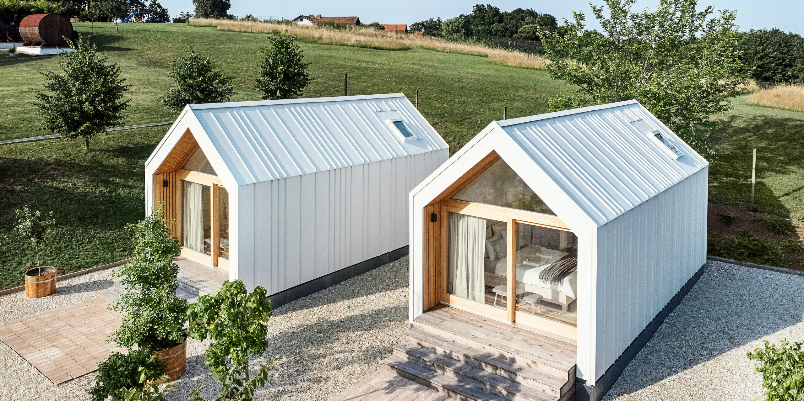 Blick auf die Vorderseite der beiden Tiny Häuser der Ferienanlage 'Pri Momi' in Kančevci, Slowenien. Die Mikrohäuser wurden in einer Holzrahmenbauweise erreichtet und mit einer robusten PREFALZ Dach- und Fassadenverkleidung in P.10 Reinweiß vollendet. Die modernen, minimalistischen Bauwerke kombinieren eine langlebige und zugleich ästhetische Aluminiumhülle mit großen Glasfronten und naturbelassenem Holz, eingebettet in eine grüne Landschaft. Die großzügigen Holzstufen führen zu den Eingängen, die eine gemütliche und einladende Atmosphäre schaffen.