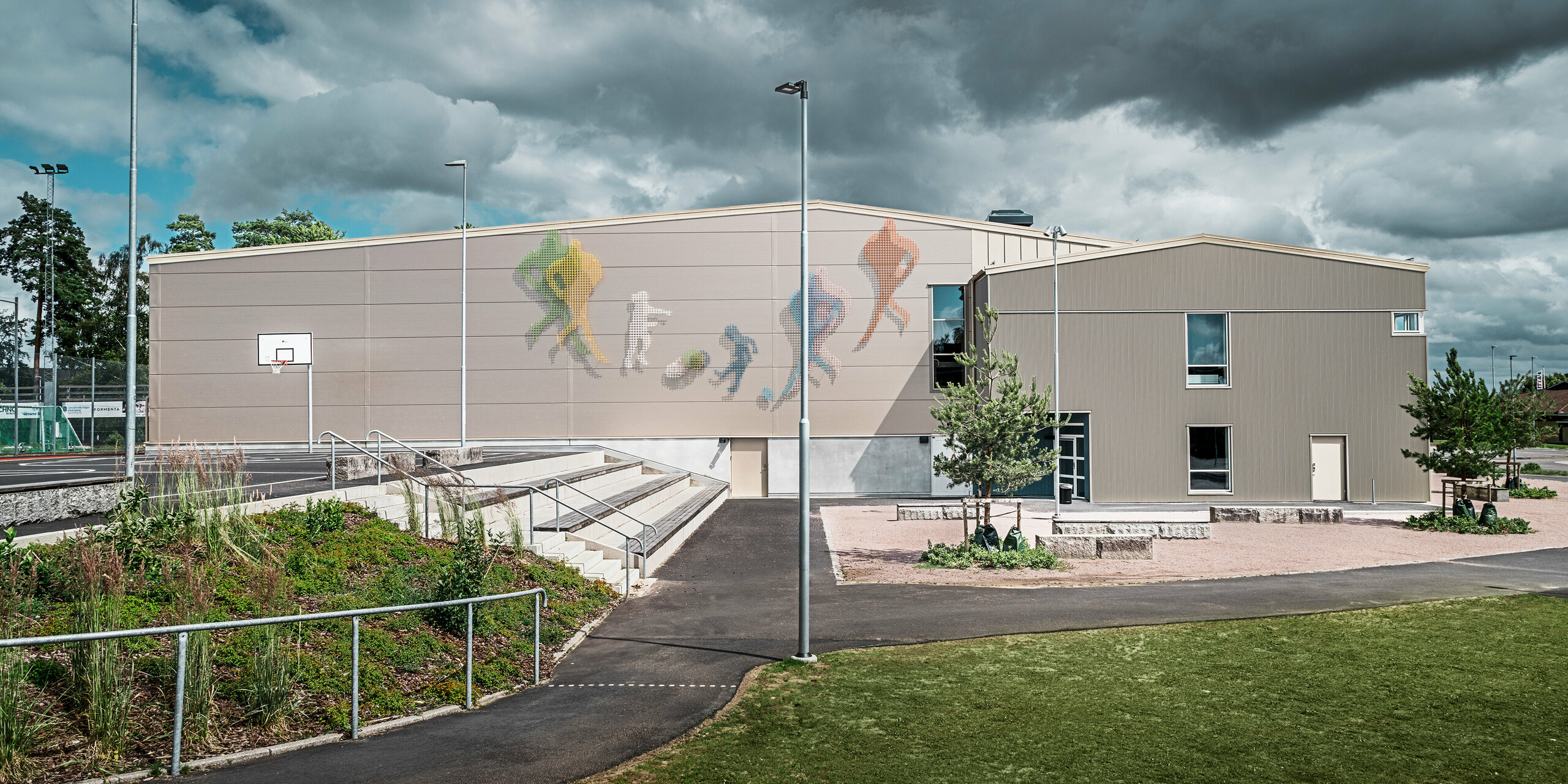 Vue extérieure du Park Arena de Värnamo, en Suède, avec une œuvre d'art murale colorée représentant des personnages sportifs. La façade est revêtue du profil triangle PREFA en bronze, qui assure une enveloppe de bâtiment moderne et durable. Au premier plan, on distingue l'aire de repos et un terrain de basket-ball, qui soulignent le caractère sportif de l'installation.