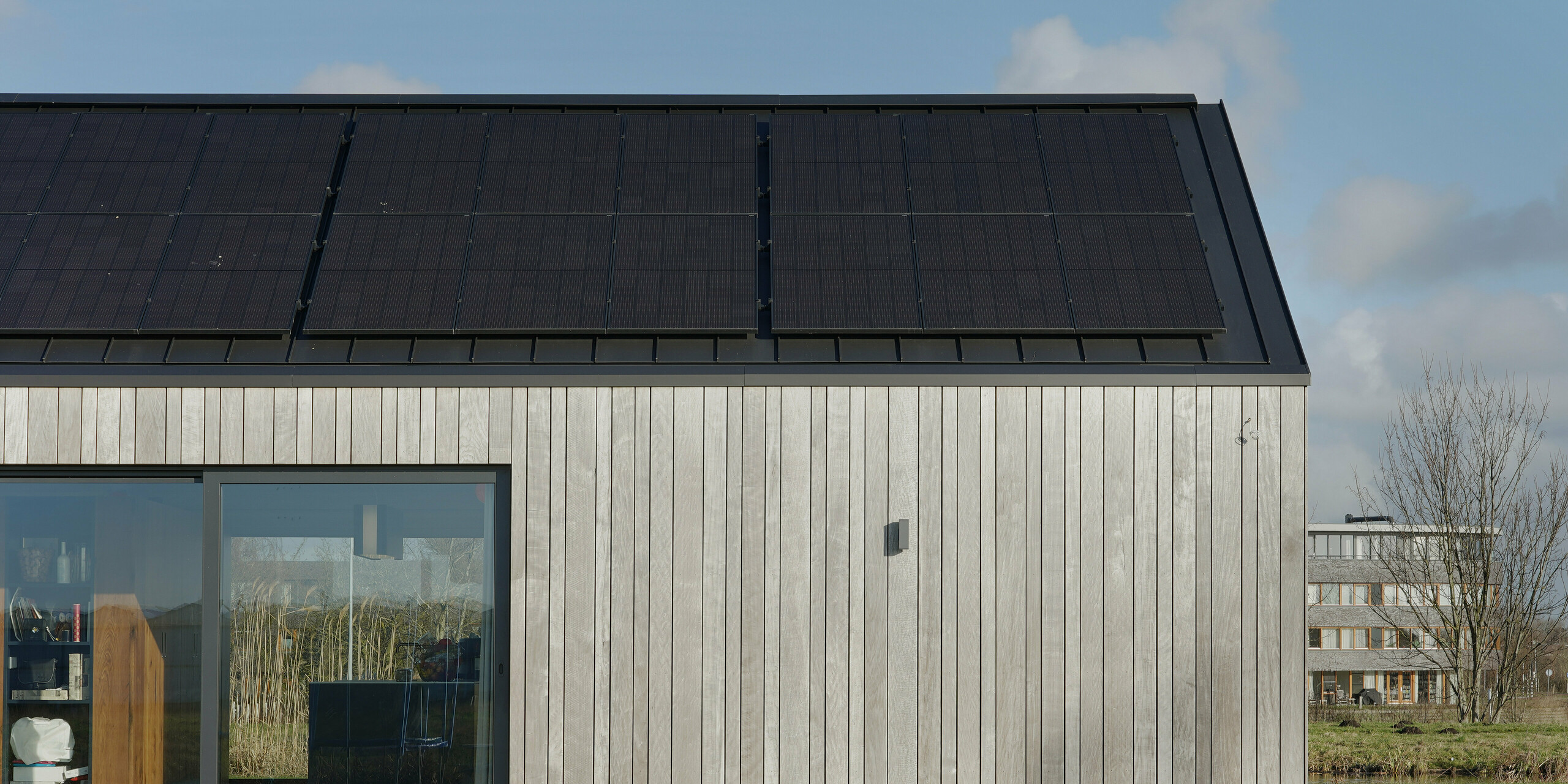 Vue détaillée d'une maison familiale moderne à Uithoorn, aux Pays-Bas, avec toiture à joints debout PREFALZ en gris noir et système photovoltaïque intégré. La photo montre l'intégration précise du système photovoltaïque sur le toit en aluminium de haute qualité ainsi que les éléments en bois naturel sur la façade. La combinaison de matériaux durables et de lignes claires souligne la construction durable et résistante aux intempéries. Idéal pour les projets de construction innovants qui reposent sur les énergies renouvelables et un design moderne. Le bungalow à l'architecture attrayante dispose également de généreuses fenêtres qui créent une atmosphère de vie invitante.