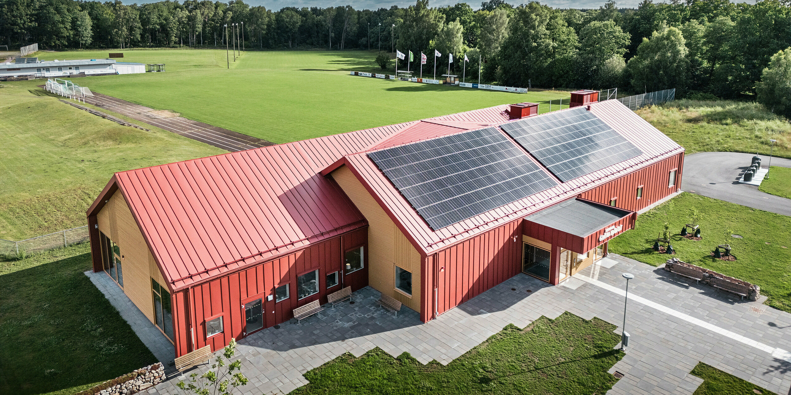 Vue aérienne de la cantine scolaire Restaurang Gröna Hjärtat à Broby, en Suède, montrant le bâtiment avec une toiture à joints debout remarquable et la façade en PREFALZ en rouge oxyde. Un système photovoltaïque à grande échelle est installé sur le toit, qui fournit de l'électricité à partir d'énergies renouvelables pour le bâtiment. Les lignes architecturales claires sont soulignées par la combinaison contrastée de l'aluminium rouge et du bois chaleureux sur les pignons. En arrière-plan se trouve un grand terrain de sport intégré dans un cadre verdoyant. Ce cliché met l'accent sur l'intégration harmonieuse du bâtiment moderne dans le paysage naturel.