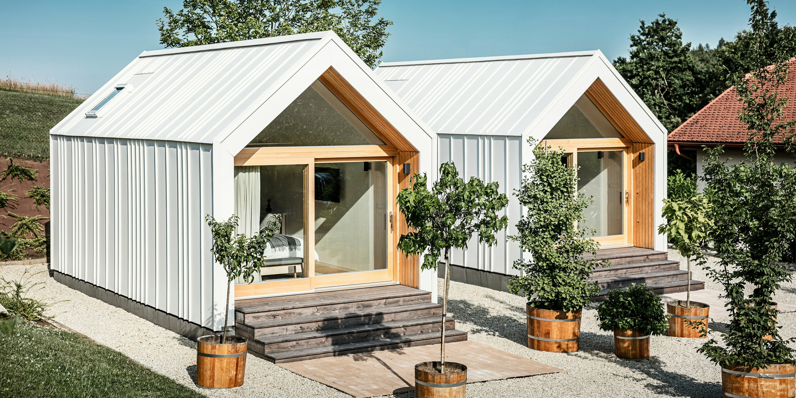 Vue de face des deux petites maisons du complexe de vacances « Pri Momi » à Kančevci, en Slovénie. L'hébergement exclusif pour les invités a été recouvert d'un toit et d'un revêtement de façade robustes PREFALZ dans la couleur P.10 blanc pur. Les micro-maisons modernes combinent une coque en aluminium esthétique et durable avec de grandes façades vitrées et du bois naturel, créant une atmosphère invitante et chaleureuse. Les environs sont caractérisés par des pelouses bien entretenues et de petits arbres dans des jardinières qui complètent le design minimaliste des maisons.