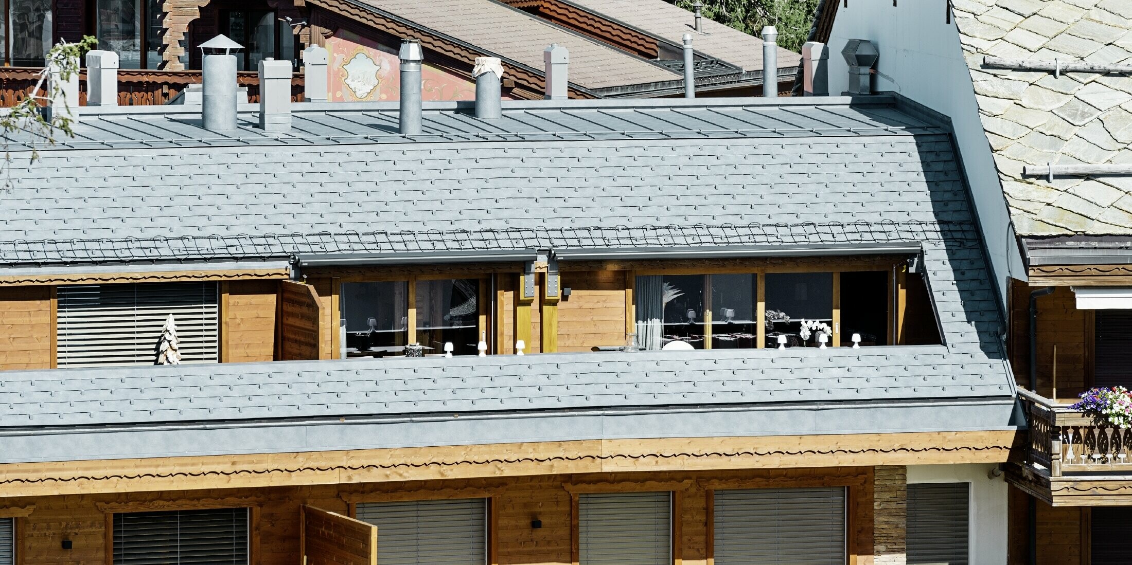 Mehrfamilienhaus in Crans-Montana mit Bergen im Hintergrund und einer Fassade mit verspielten Holzelementen mit einem PREFA Aluminium Dachschindel-Dach in steingrau 