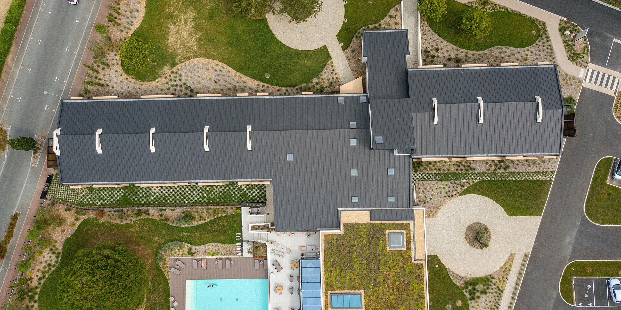 Prise de vue holistique par drone de l'Hôtel Parc du Landreau aux Herbiers, France, avec toit PREFALZ en gris noir. Le toit en joint debout en aluminium durable offre une protection optimale contre les intempéries et s'intègre harmonieusement dans le concept architectural de l'hôtel. L'espace extérieur avec piscine et toit végétalisé témoigne de la construction moderne et durable.