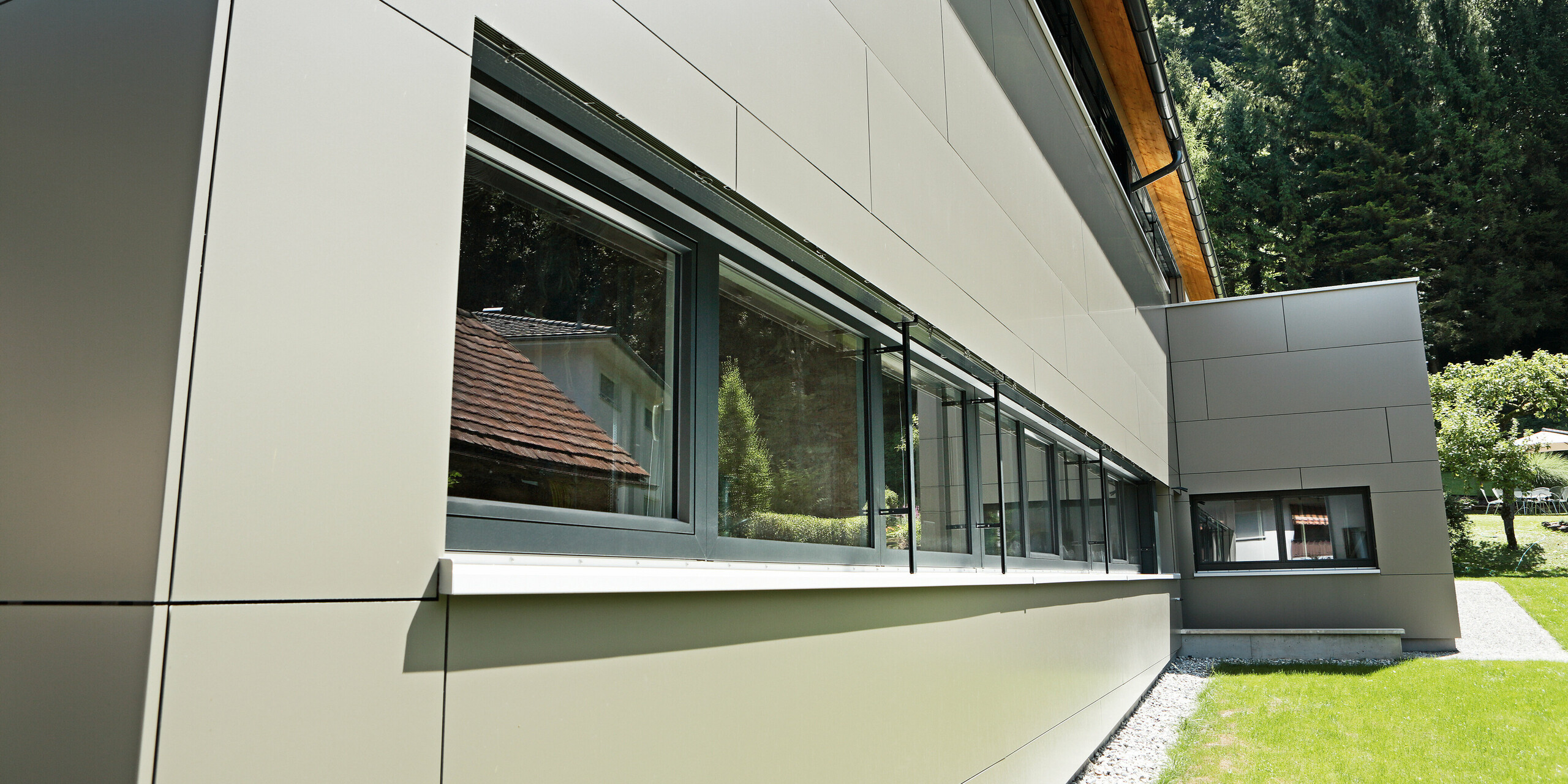 Nahaufnahme des Erdgeschosses eines Einfamilienhauses in Hohenems, das mit PREFA Aluminium Verbundplatten in der Farbe Bronzemetallic verkleidet ist. Die glatten, sauber verarbeiteten Fassadenelemente bieten eine elegante Optik und unterstreichen die großen Fensterpartien, die das Tageslicht maximieren. Die Detailaufnahme zeigt die präzise Handwerkskunst und Qualität der Materialien, die das Haus mit einem zeitgemäßen und langlebigen Äußeren versehen.