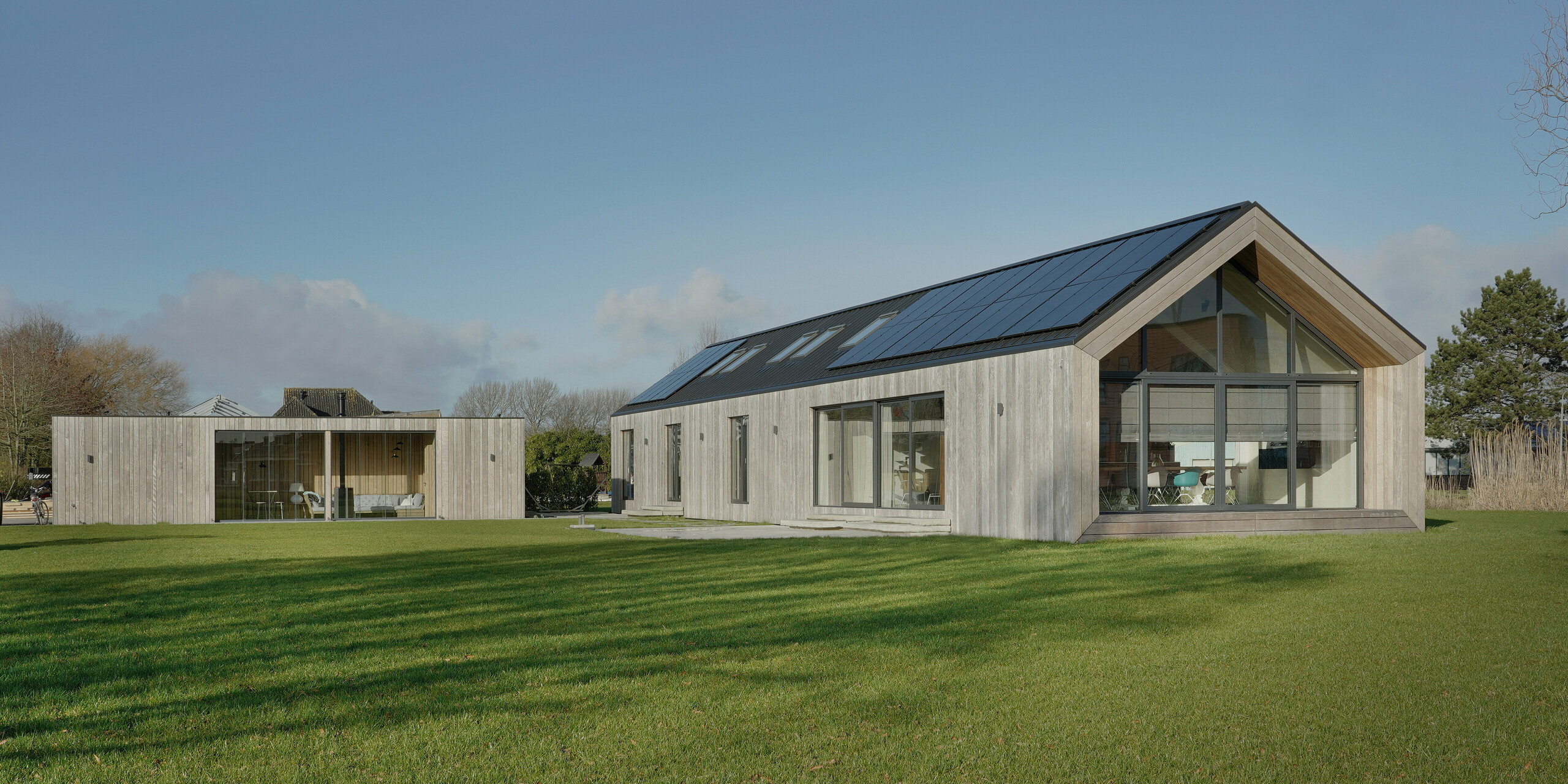 Prise de vue grand angle d'une maison familiale moderne à Uithoorn, aux Pays-Bas, avec une toiture à joints debout PREFALZ en gris noir et un système photovoltaïque intégré. La photo montre l'ensemble de la propriété avec un jardin spacieux et une combinaison de grandes façades vitrées et d'éléments en bois naturel sur la façade. Les lignes claires et les matériaux de haute qualité résistants aux intempéries soulignent la durabilité et le design moderne de la maison. Le bungalow d'un étage a été construit pour répondre aux besoins de vie d'une grande famille.