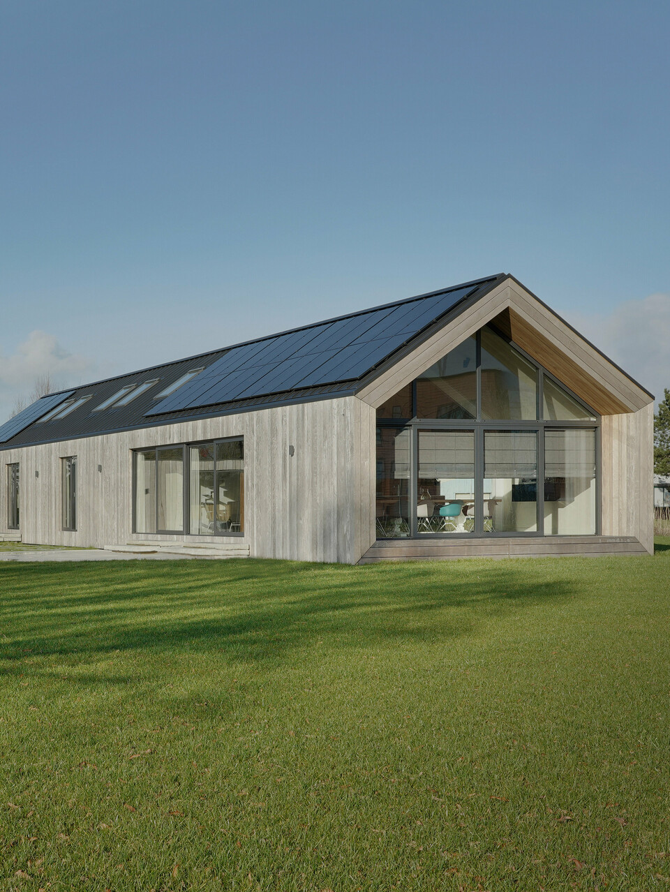 Prise de vue grand angle d'une maison familiale moderne à Uithoorn, aux Pays-Bas, avec une toiture à joints debout PREFALZ en gris noir et un système photovoltaïque intégré. La photo montre l'ensemble de la propriété avec un jardin spacieux et une combinaison de grandes façades vitrées et d'éléments en bois naturel sur la façade. Les lignes claires et les matériaux de haute qualité résistants aux intempéries soulignent la durabilité et le design moderne de la maison. Le bungalow d'un étage a été construit pour répondre aux besoins de vie d'une grande famille.