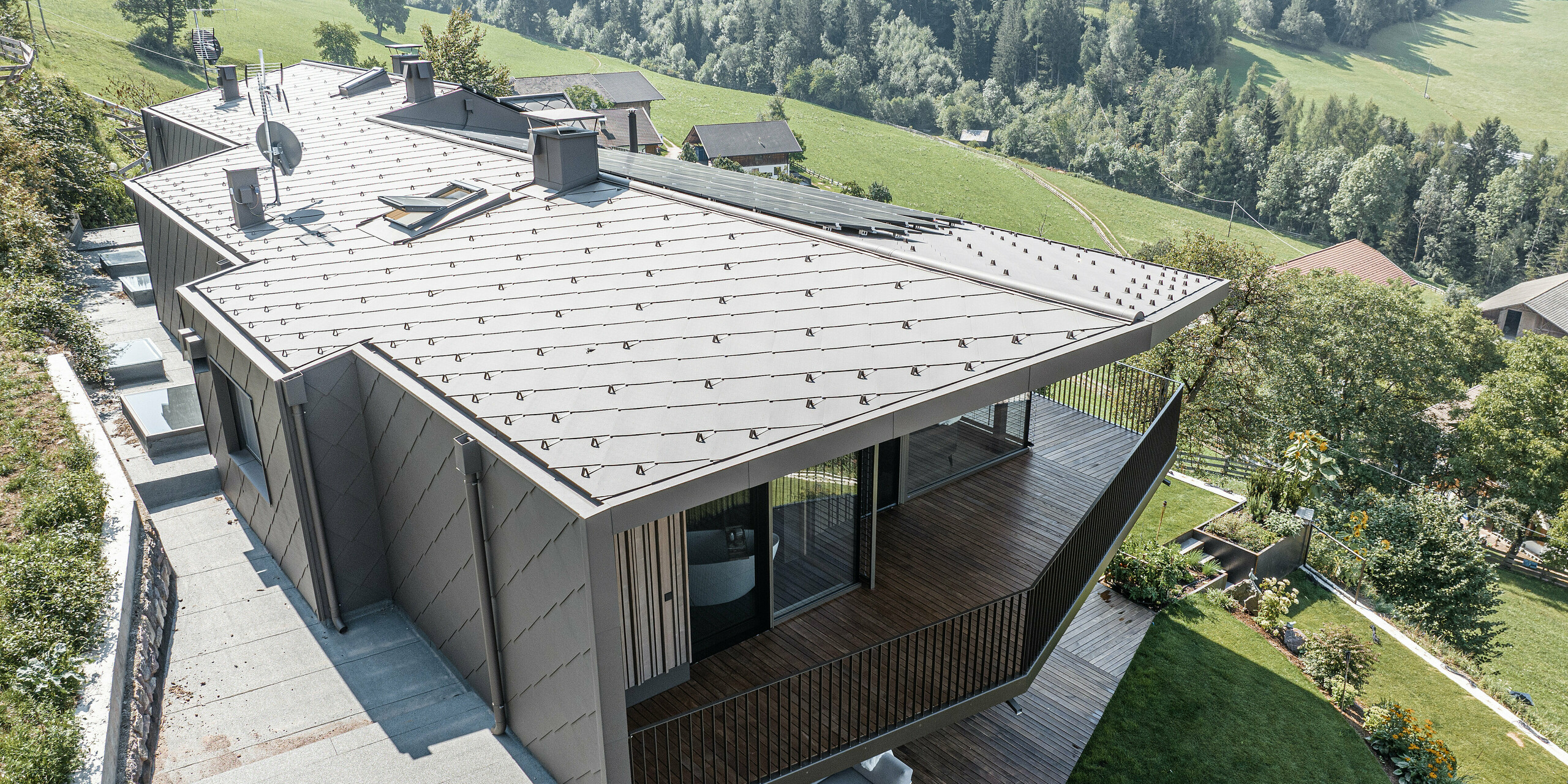 Pohled na zmodernizovaný dvojdům v Möltenu v Jižním Tyrolsku vybavený odolnými falcovanými a falcovanými šablonami 44x44 v barvě P.10 tmavě hnědé. Design střechy a fasády kombinuje robustní hliník s dřevěnými prvky, obklopený malebnou krajinou s rozsáhlými výhledy na zalesněné kopce. Redesign plynule integruje zrekonstruovanou stávající budovu a nově vybudovaný areál do harmonického celku se zaměřením na dřevo jako stavební materiál.
