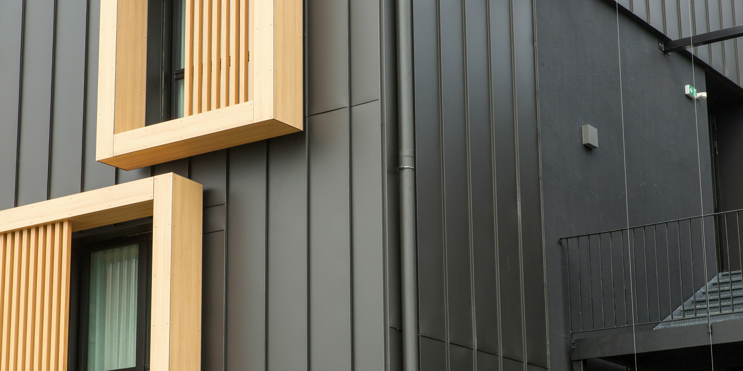 Vue détaillée de la façade de l'Hôtel Parc du Landreau aux Herbiers, France, avec un revêtement en aluminium de PREFA. La façade gris noir PREFALZ est complétée par le système d'évacuation des eaux pluviales PREFA intégré. Le tuyau de descente présenté s'intègre harmonieusement dans le système de façade et assure un drainage efficace et durable des eaux de pluie. La combinaison du bois et du revêtement à joint debout de haute qualité en tôle souligne le design moderne. Sur le bord droit de l'image, vous pouvez voir l'escalier harmonieusement intégré dans la même palette de couleurs.