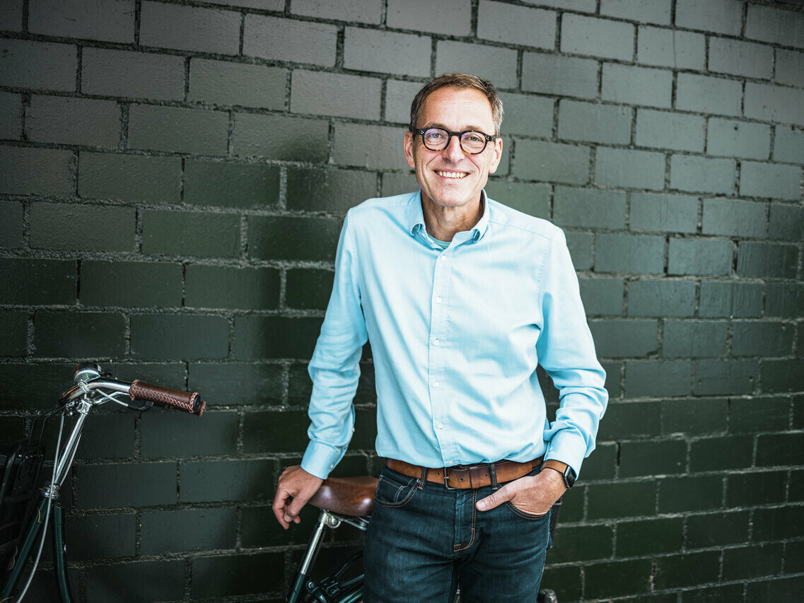 Portrait des Architekten Jochen Weissenrieder, lehnt an einem Fahrrad, im Hintergrund eine schwarz-graue Ziegelwand.