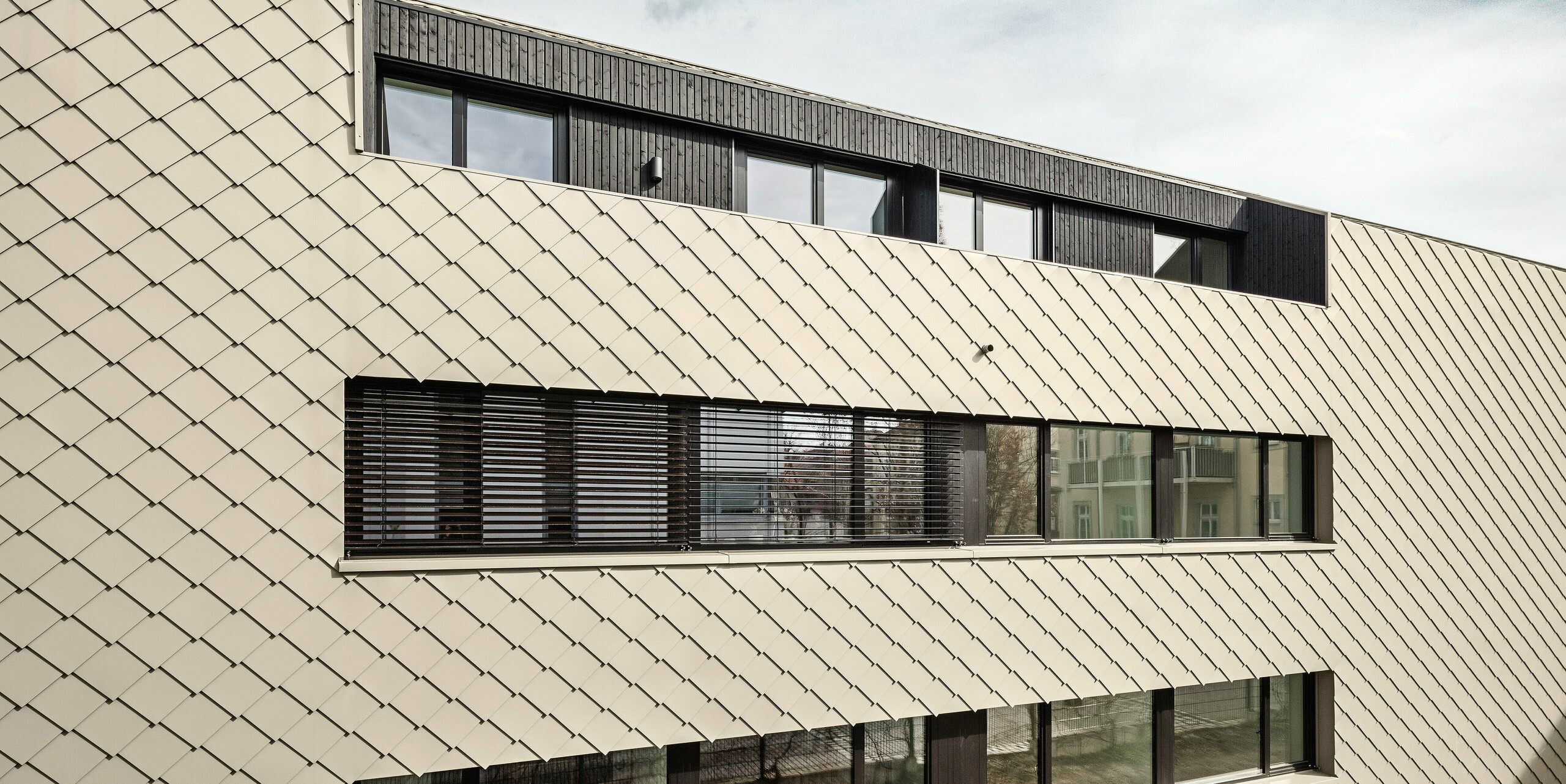 Vue arrière d'un immeuble d'appartements moderne à Dresde-Plauen avec une façade en losanges PREFA 29 × 29 en bronze. La photo montre le revêtement de façade à structure uniforme avec de grandes fenêtres disposées symétriquement qui laissent entrer beaucoup de lumière naturelle à l'intérieur. La combinaison du revêtement de couleur bronze et des encadrements de fenêtres sombres souligne la construction bien pensée du bâtiment.