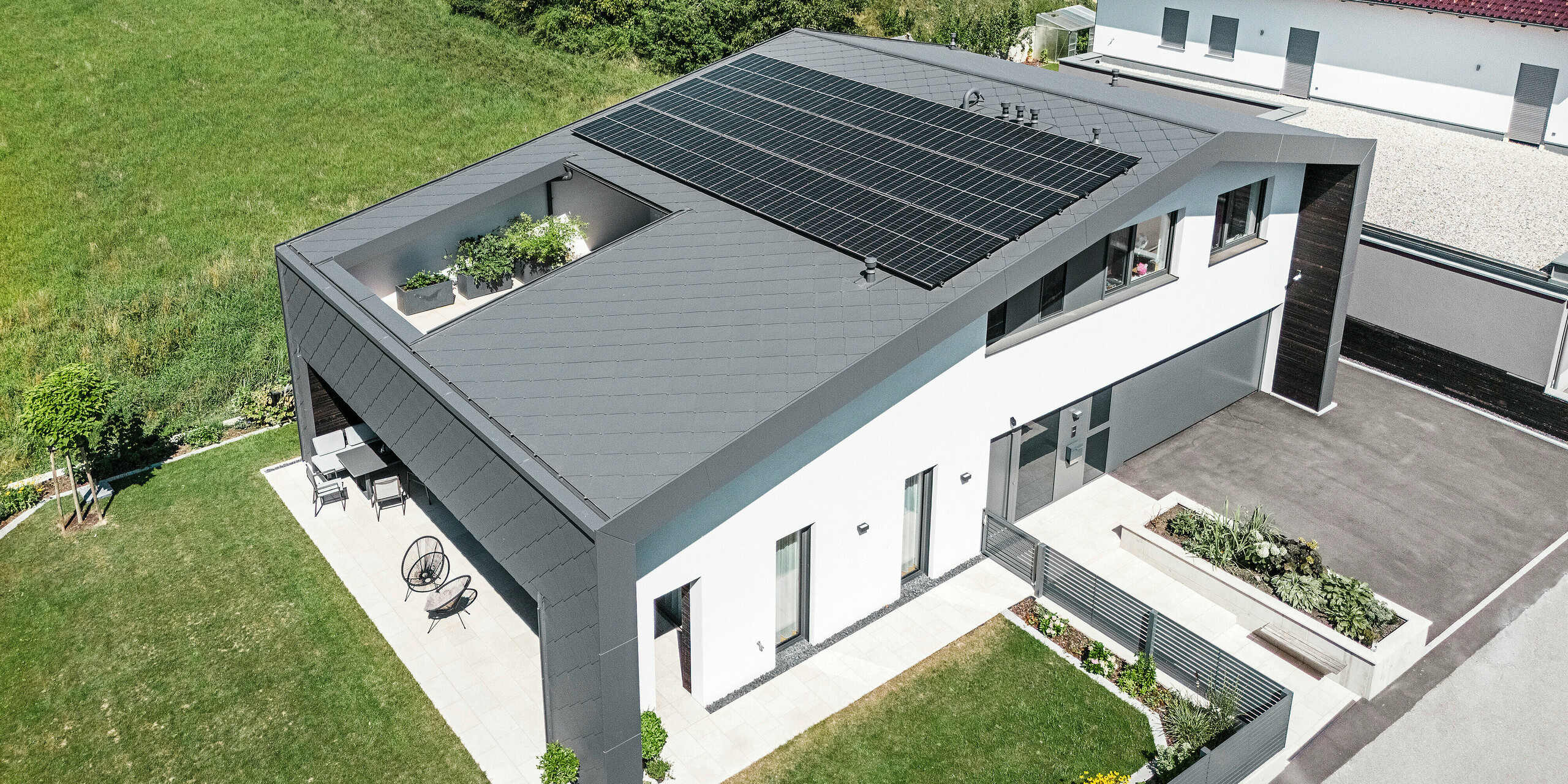 Prise de vue par drone d'une maison familiale moderne à Aschach avec des losanges de toiture et de façade 44x44 gris sombre  dans le revêtement P.10 de haute qualité. Le toit spacieux est équipé d'un système photovoltaïque et d'un atrium. L'architecture attrayante intègre également une terrasse couverte. Le toit et la façade en aluminium sont légers, durables et offrent une protection optimale contre les intempéries. La façade du bâtiment est en crépi blanc.