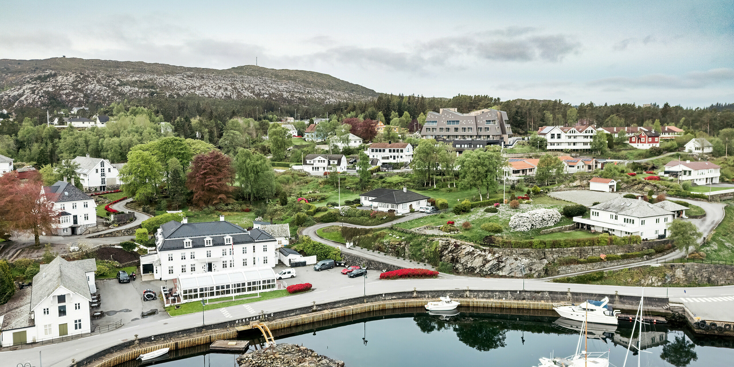 Vue aérienne du port de Bekkjarvik, en Norvège, avec vue sur la maison Beckerwyc en arrière-plan. L'hôtel se distingue des maisons traditionnelles en bois blanc environnantes par son architecture futuriste et sa façade en losanges de façade PREFA 44 × 44 de couleur brun noisette. Le paysage pittoresque, le port avec ses bateaux et les environs verdoyants soulignent l'emplacement unique de l'hôtel dans cette ville côtière norvégienne idyllique sur l'île de Selbjørn.