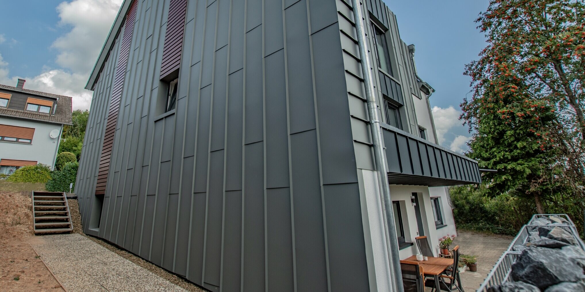 Fassadengestaltung mit vertikaler Winkelstehfalzfassade in hellgrau und Fassadenpaneelen, horizontal verlegt, in Holz dunkel. Die gesamte Fassade wurde aus Aluminium von PREFA gefertigt.