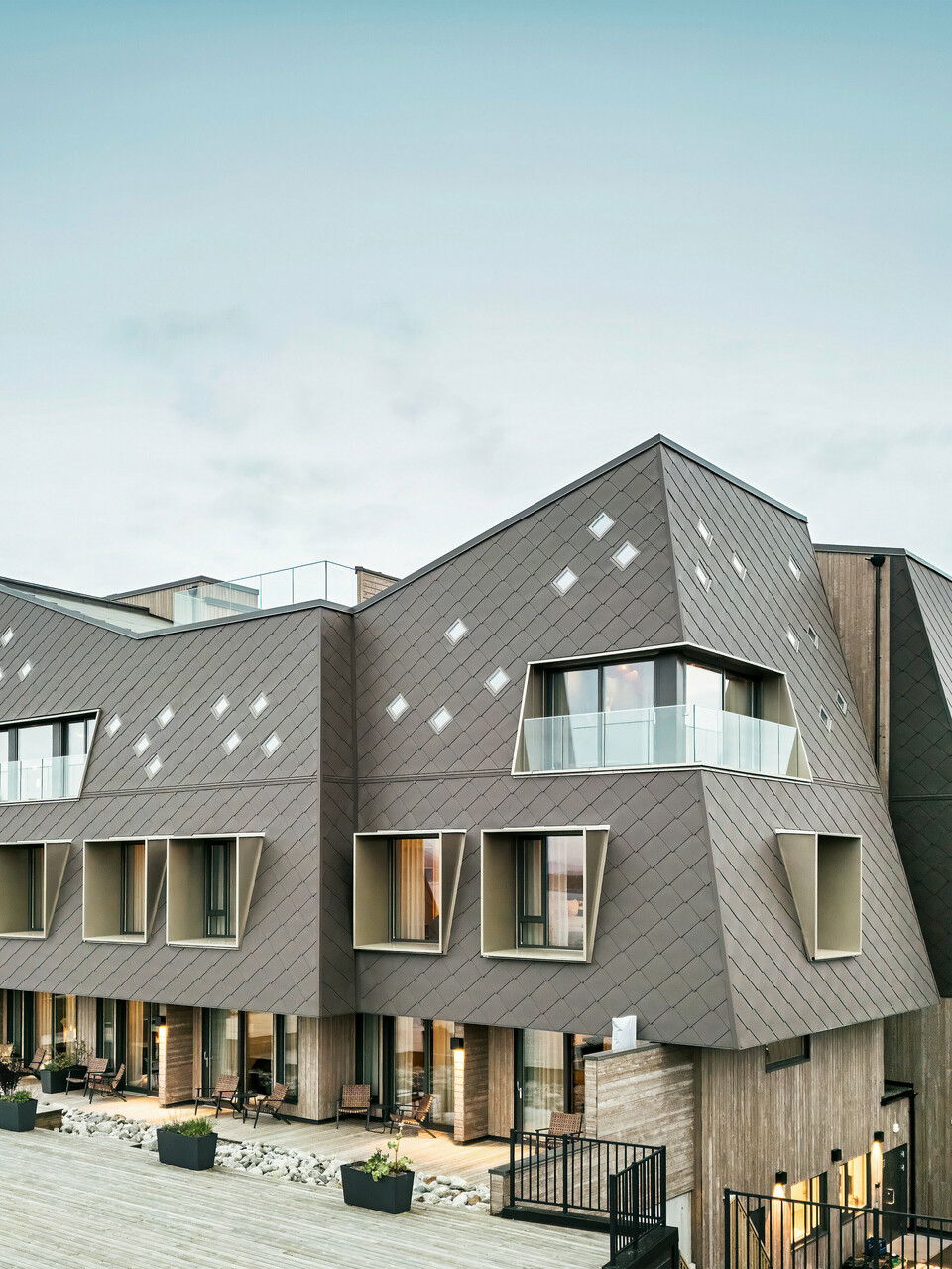 Vue de la terrasse de l'hôtel Beckerwyc House à Bekkjarvik, Norvège. Le bâtiment moderne est décoré d'une façade en aluminium frappante composée de losanges PREFA 44 × 44 en brun noisette. L'architecture futuriste avec des surfaces de toit en pente et des éléments de fenêtre proéminents est complétée par des espaces extérieurs spacieux. La couleur naturelle de la façade s'intègre harmonieusement dans le paysage environnant et souligne le design contemporain du bâtiment.