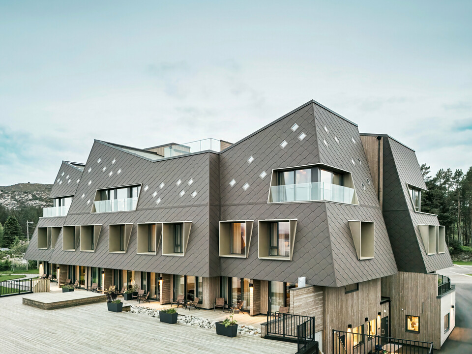 Vue de la terrasse de l'hôtel Beckerwyc House à Bekkjarvik, Norvège. Le bâtiment moderne est décoré d'une façade en aluminium frappante composée de losanges PREFA 44 × 44 en brun noisette. L'architecture futuriste avec des surfaces de toit en pente et des éléments de fenêtre proéminents est complétée par des espaces extérieurs spacieux. La couleur naturelle de la façade s'intègre harmonieusement dans le paysage environnant et souligne le design contemporain du bâtiment.