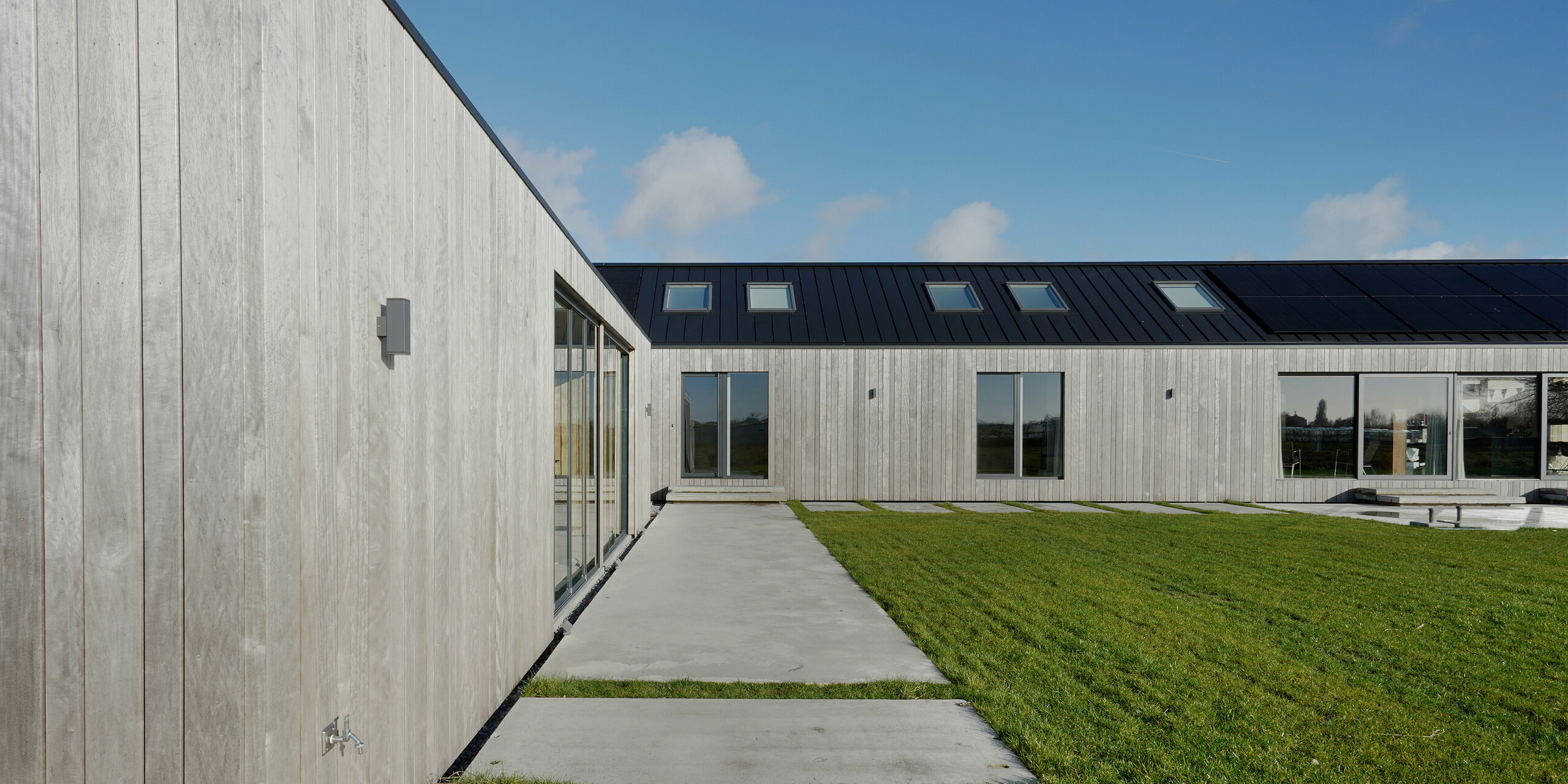 Vue latérale d'une maison familiale moderne à Uithoorn, Pays-Bas. L'image met en valeur la façade en bois naturel et le toit à joints debout en PREFALZ de couleur gris noir, conçu avec précision. L'allée pavée le long de la façade en bois et les grandes façades vitrées de la maison sont également au centre de la photographie. Les lignes claires et l'utilisation d'aluminium durable et résistant aux intempéries et de bois durable soulignent le design écologique et moderne du bâtiment. Idéal pour les projets de construction respectueux de l’environnement qui mettent l’accent sur la qualité et l’esthétique.