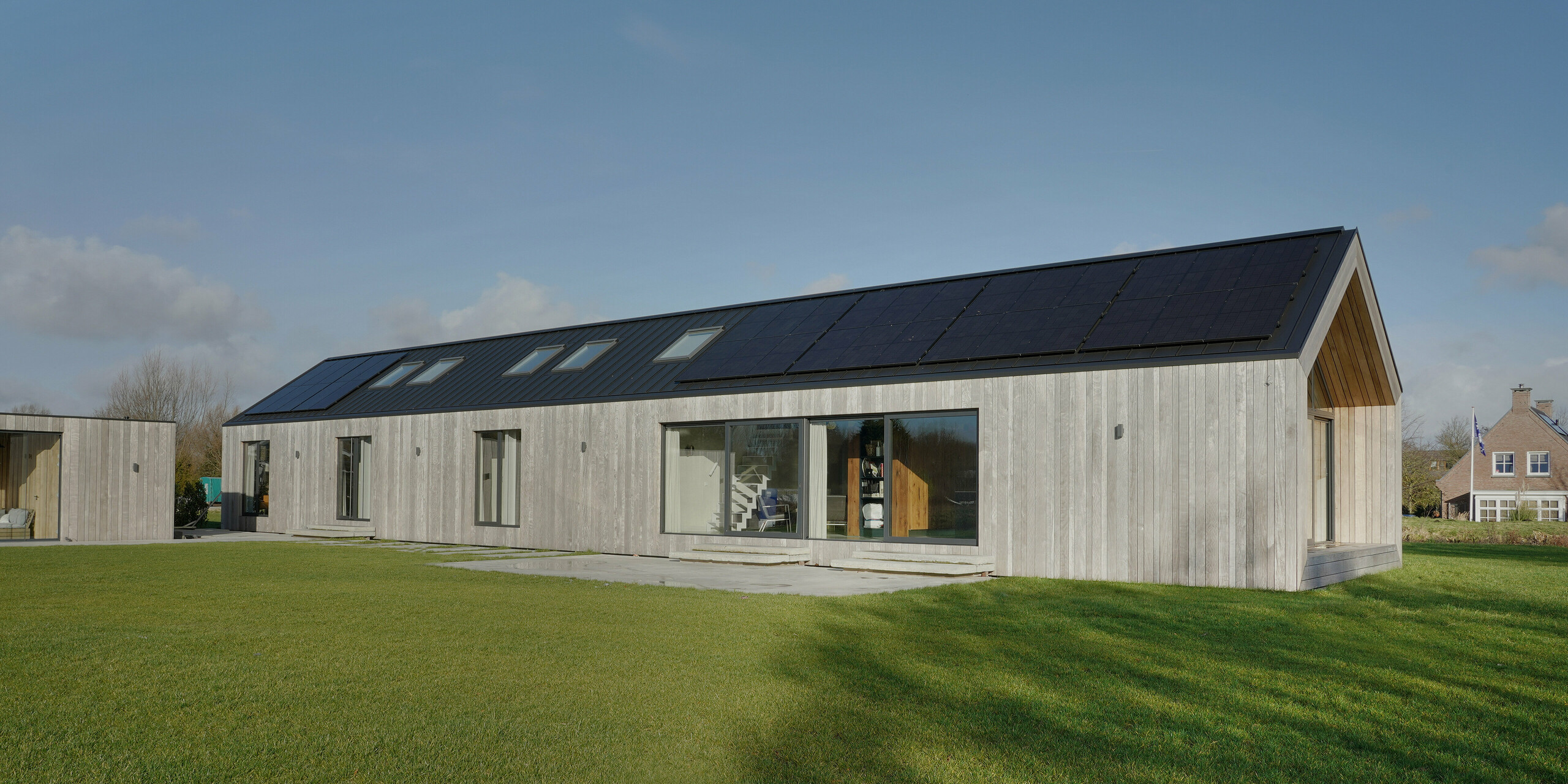 Vue grand angle d'un bungalow moderne à Uithoorn, aux Pays-Bas, avec une toiture à joints debout PREFALZ en gris noir et un système photovoltaïque comme solution de toit. La photo montre le bâtiment spacieux avec de grandes façades vitrées et un revêtement en bois, entouré d'une pelouse bien entretenue. La construction durable combine une conception esthétique avec des matériaux durables et résistants aux intempéries, idéale pour les projets de construction respectueux de l'environnement.