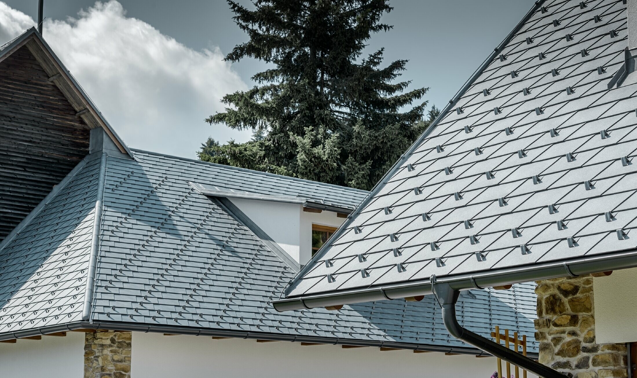 Vue détaillée d’une couverture de toit en alu de PREFA ; bardeau de toiture couleur gris pierre avec la gouttière en alu couleur anthracite de PREFA ; en arrière plan on peut voir une lucarne rampante avec toit à joint debout. La façade est blanche, avec des éléments en pierre incorporés.