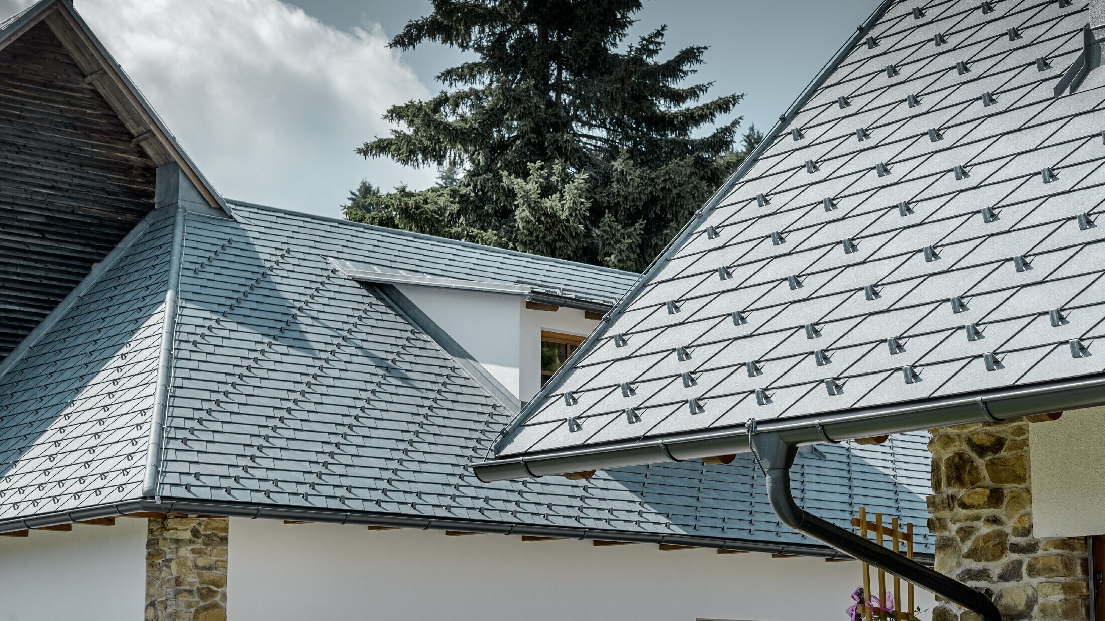 Vue détaillée d’une couverture de toit en alu de PREFA ; bardeau de toiture couleur gris pierre avec la gouttière en alu couleur anthracite de PREFA ; en arrière plan on peut voir une lucarne rampante avec toit à joint debout. La façade est blanche, avec des éléments en pierre incorporés.