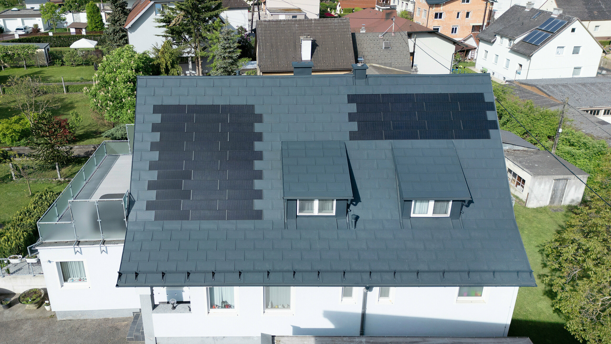 Sanierung eines Einfamilienhauses in Leonding mit PREFA Solardachplatten in Anthrazit. Das Bild zeigt das Haus aus der Vogelperspektive nach der Dachsanierung. Integrierte Solarmodule auf dem Dach bieten eine nachhaltige Energiequelle und sind nahtlos in die Dachfläche eingebettet. Das Projekt demonstriert die innovative Kombination von Dachsanierung und Photovoltaiksystemen mit langlebigen und witterungsbeständigen PREFA Materialien. Ideal für nachhaltiges Bauen und energieeffiziente Lösungen.