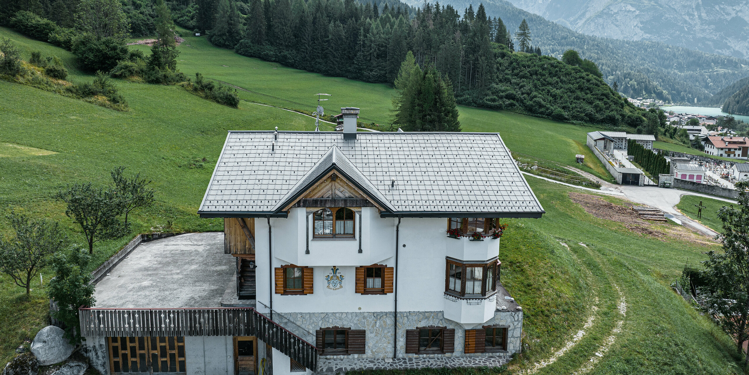 Pohled z ptačí perspektivy na vchod do tradiční horské chaty v Auronzo di Cadore po rekonstrukci střechy, vyznačující se falcovanými taškami PREFA v barvě P.10 světle šedé. Budova s ​​charakteristickým alpským šarmem a moderní střechou je v harmonii se svěží zelenou krajinou a působivým horským panoramatem v pozadí a demonstruje úspěšnou kombinaci funkčnosti a regionální architektury.