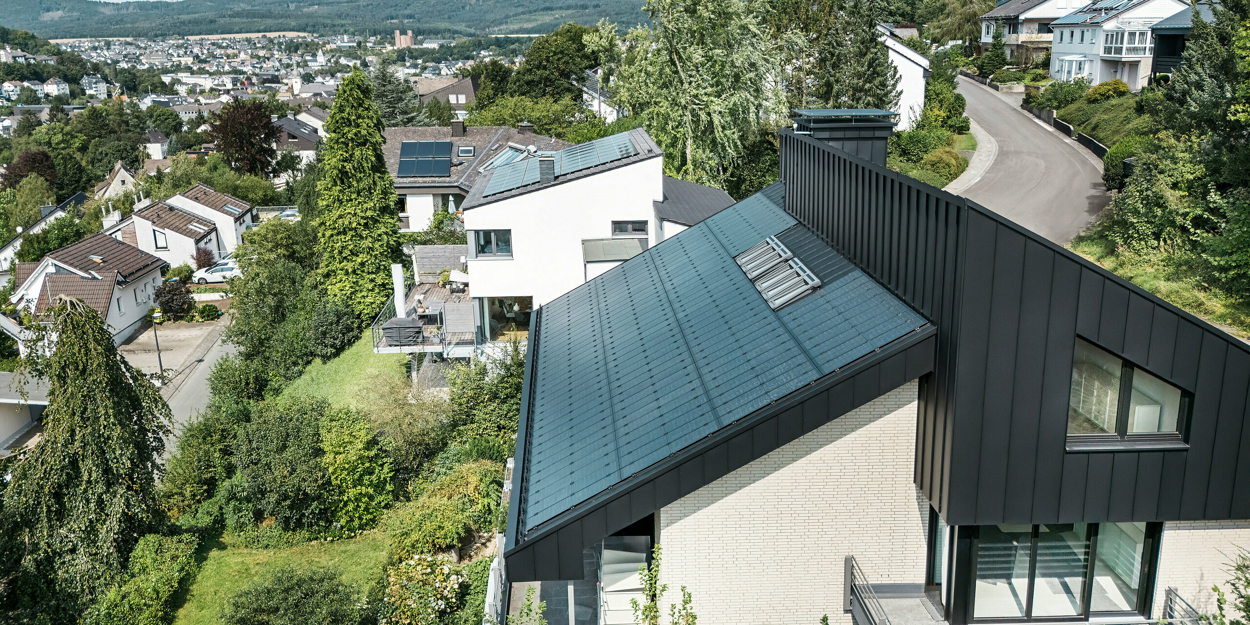 Vue panoramique d'une maison unifamiliale à Meschede, en Allemagne, avec un toit en pente décalé en PREFALZ en P.10 Noir. Le toit est équipé de modules photovoltaïques verre/verre sans cadre de PREFA, qui allient durabilité et design. Le revêtement en aluminium de la façade du pignon complète les lignes épurées et l'architecture moderne, intégrée dans un quartier résidentiel verdoyant avec vue sur le paysage environnant et la ville.