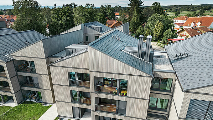 Modernes Mehrfamilienhaus in Utting am Ammersee, gekennzeichnet durch eine innovative Hybridbauweise mit einer eleganten Holzfassade und hochwertigen PREFA Dachsystemen aus Aluminium. Verwendet wurden dunkelgraue Dachplatten R.16 sowie PREFALZ. Das Gebäude besticht durch klare Linien, nachhaltige Materialien und eine harmonische Integration in die grüne Umgebung. Die architektonische Gestaltung verbindet Ästhetik und Funktionalität, wobei die langlebigen und wetterbeständigen PREFA Produkte maßgeblich zur hohen Qualität und eindrucksvollen Optik beitragen.