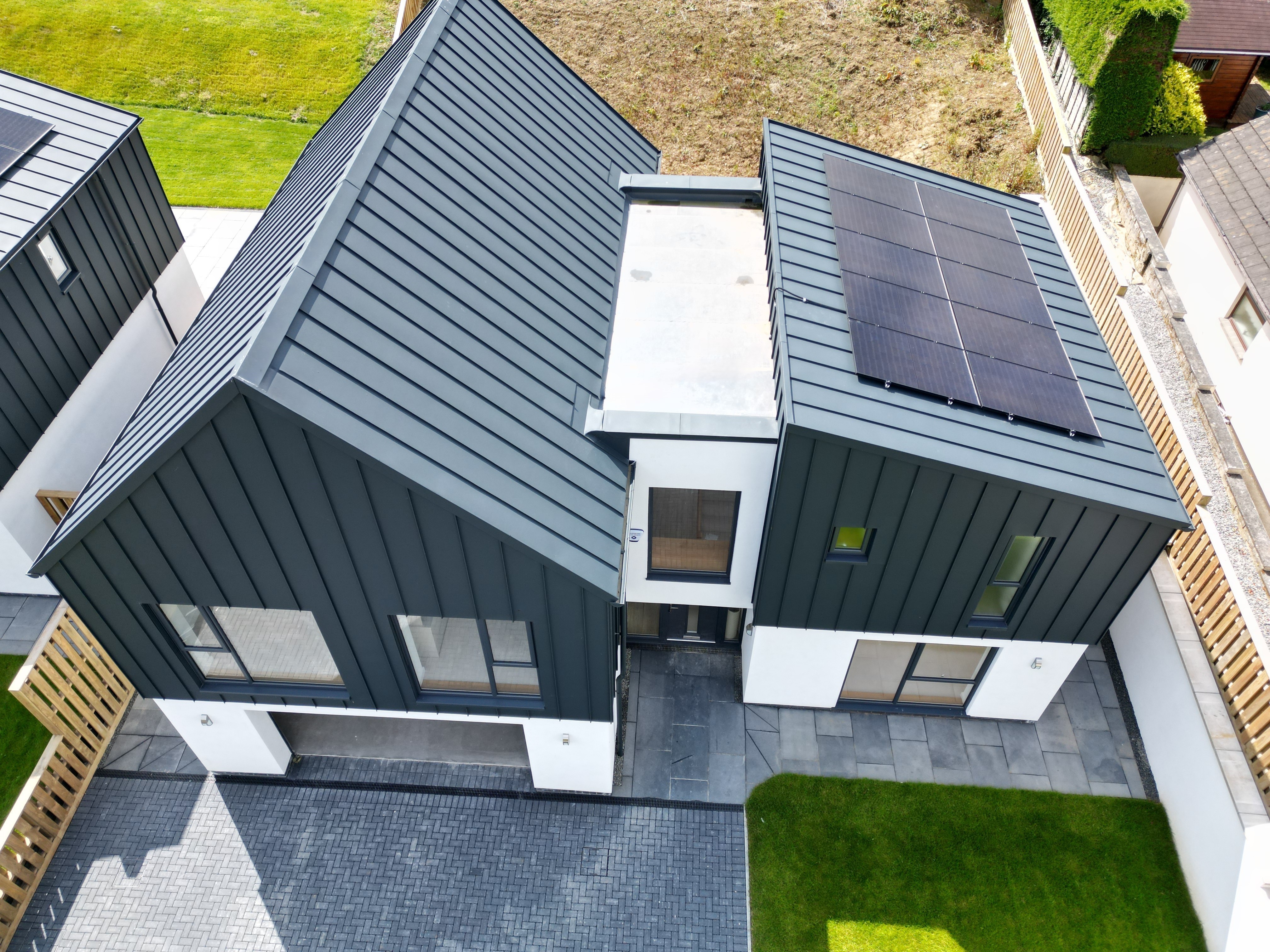 Vogelperspektive auf ein Einfamilienhaus in Bridgerule, Devon, mit PREFALZ P.10 anthrazit als Stehfalzdeckung auf Dach und Fassade. Die moderne Architektur verbindet langlebiges Aluminium mit ästhetischer Langlebigkeit, ergänzt durch eine Solaranlage. Über der Garageneinfahrt thront ein Satteldach, der rechte Gebäudeteil erhielt ein Pultdach. Zwischen den Dächern verbindet eine Gebäudebrücke die beiden Baukörper.